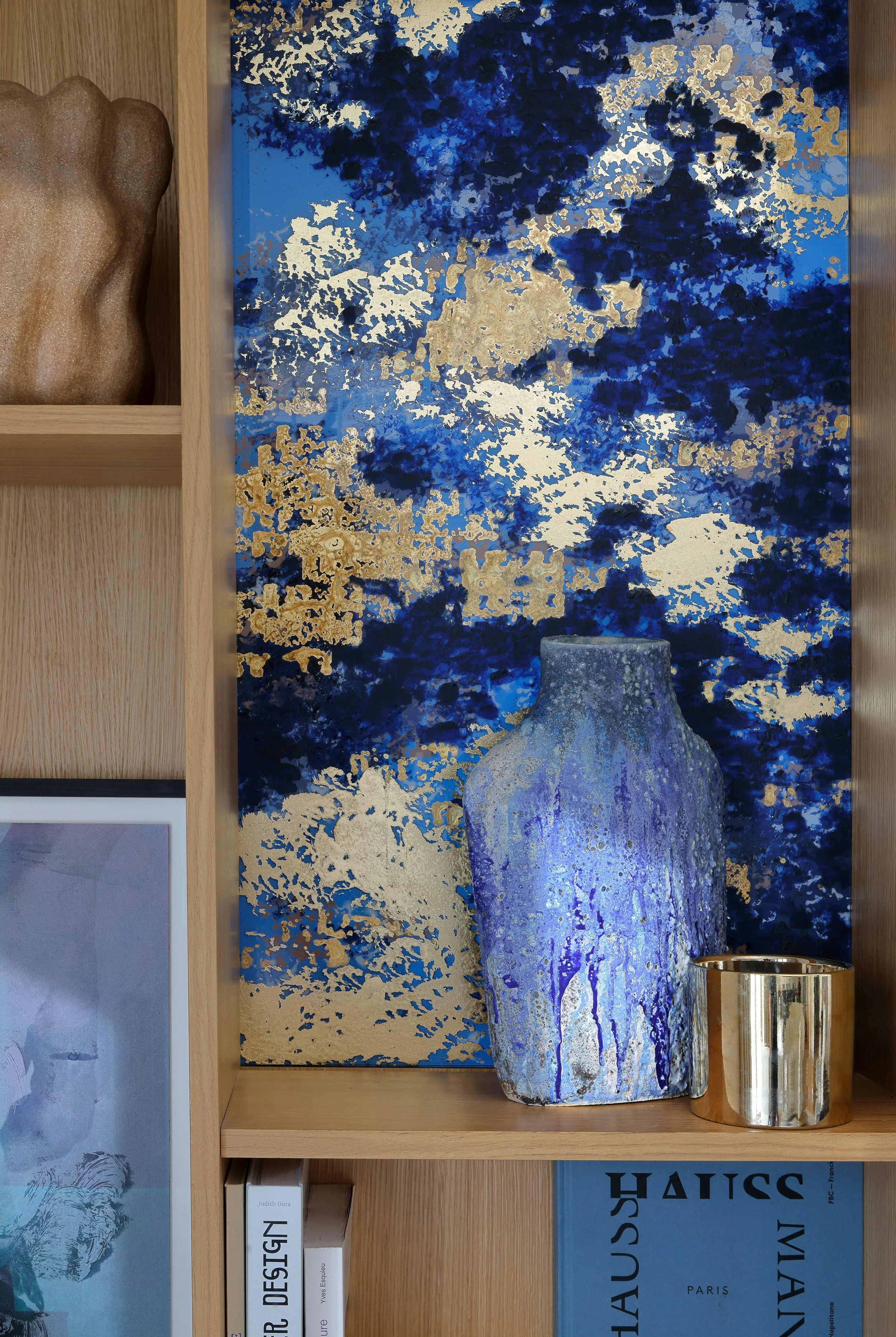 Une étagère en bois avec un grand vase en céramique bleu et violet, un petit pot en métal doré, et un tableau artistique avec des éclaboussures de bleu, d'or et de blanc en fond.