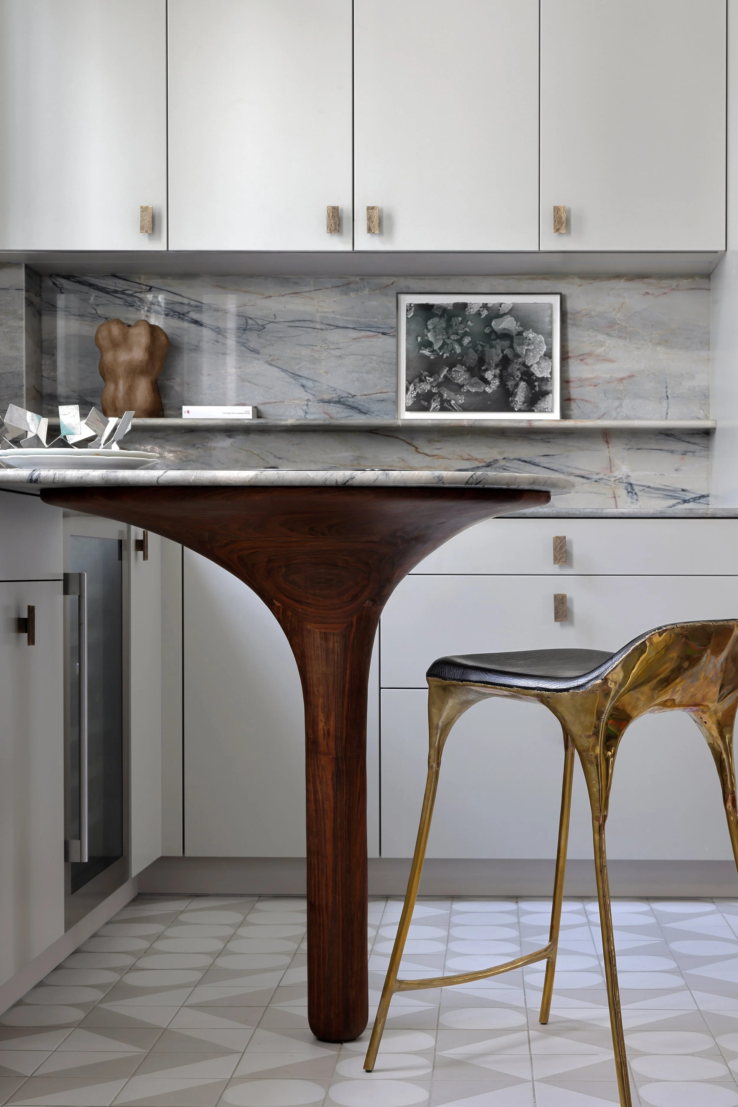 Cuisine moderne avec comptoir en marbre gris, meuble blanc, table en bois incurvée, chaise en métal doré, décoration murale, sculpture en forme de torse et une photo en noir et blanc.