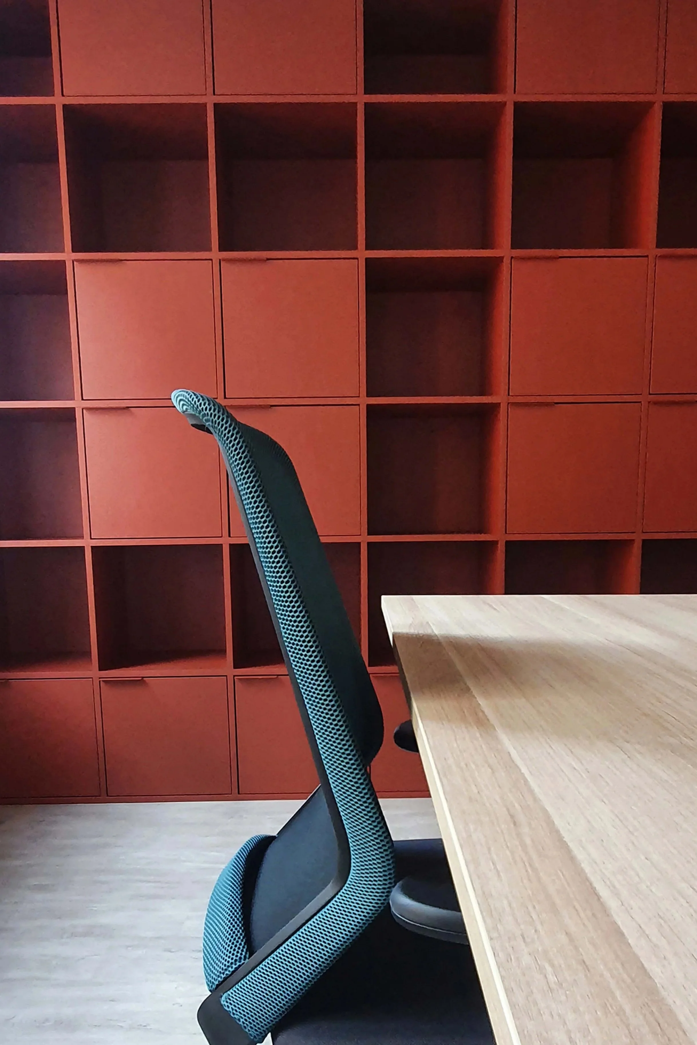 A black and blue ergonomic armchair next to a light wood table, with a square wall of red wood chests or shelves.
