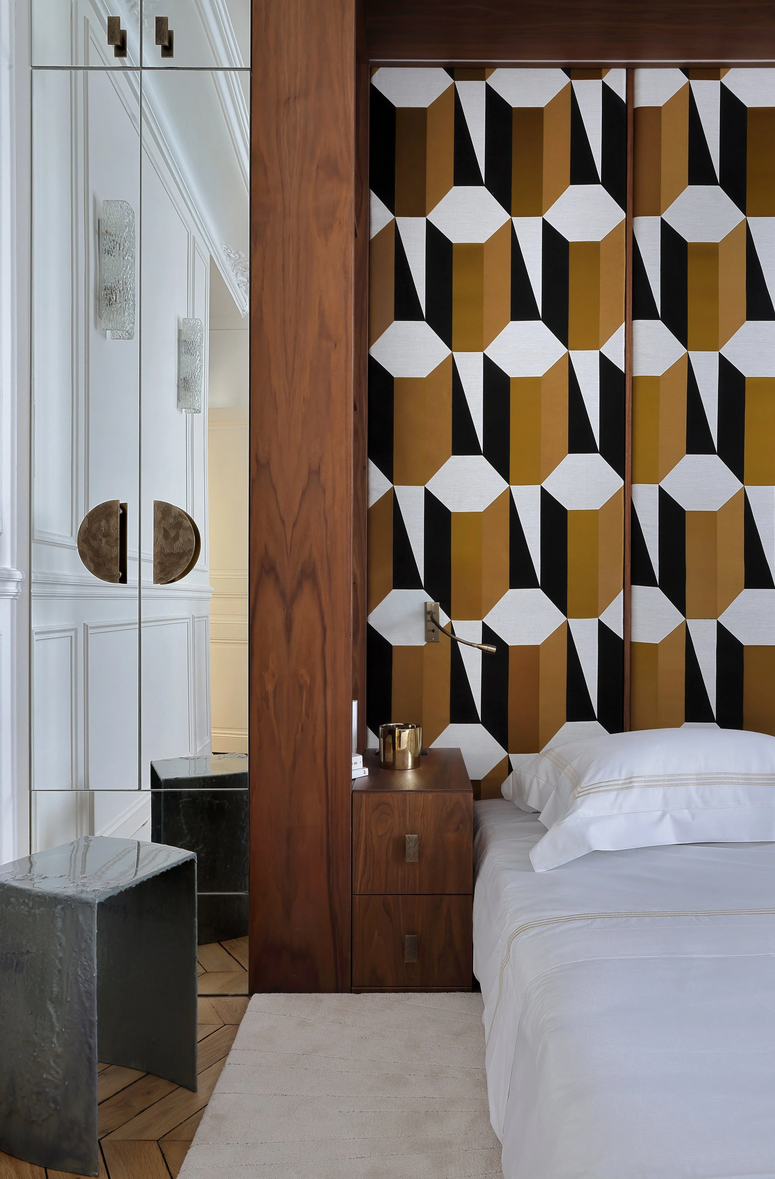 Chambre avec tête de lit en bois et mur à motif géométrique en noir, blanc et or. Côté gauche, armoire blanche avec miroir et deux petits tabourets noirs avec reflets métalliques. Côté droit, lit avec draps blancs et oreillers.