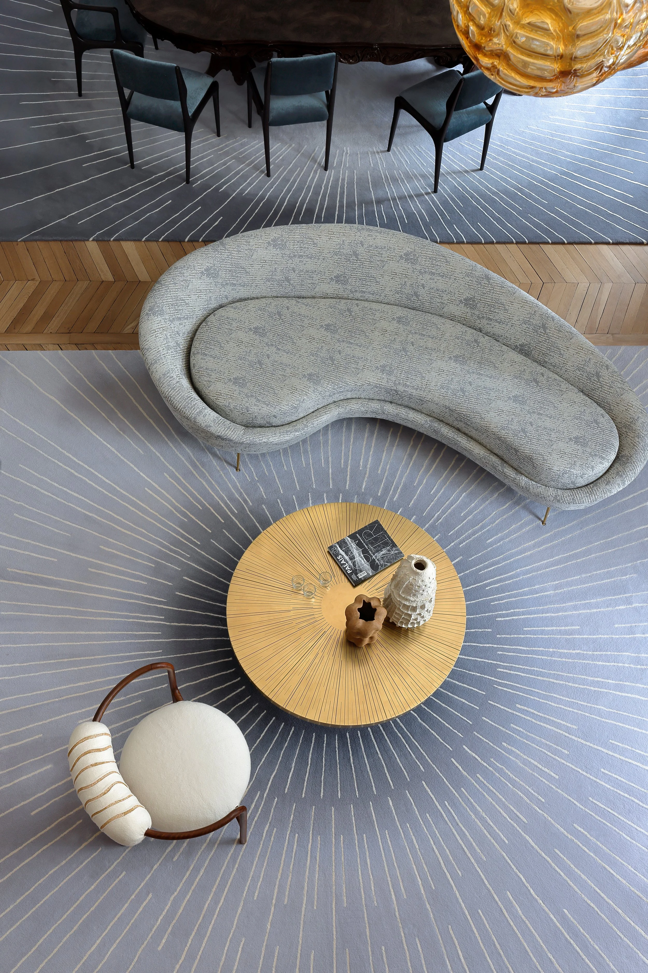 Une vue d'ensemble d'un salon ou espace de vie avec un canapé gris, une table ronde en bois clair, une chaise design blanche avec un coussin, et un tapis gris à motifs. La surface en haut de l'image montre une table ronde en bois foncé entourée de chaises bleues, avec un éclairage en suspension en verre doré.