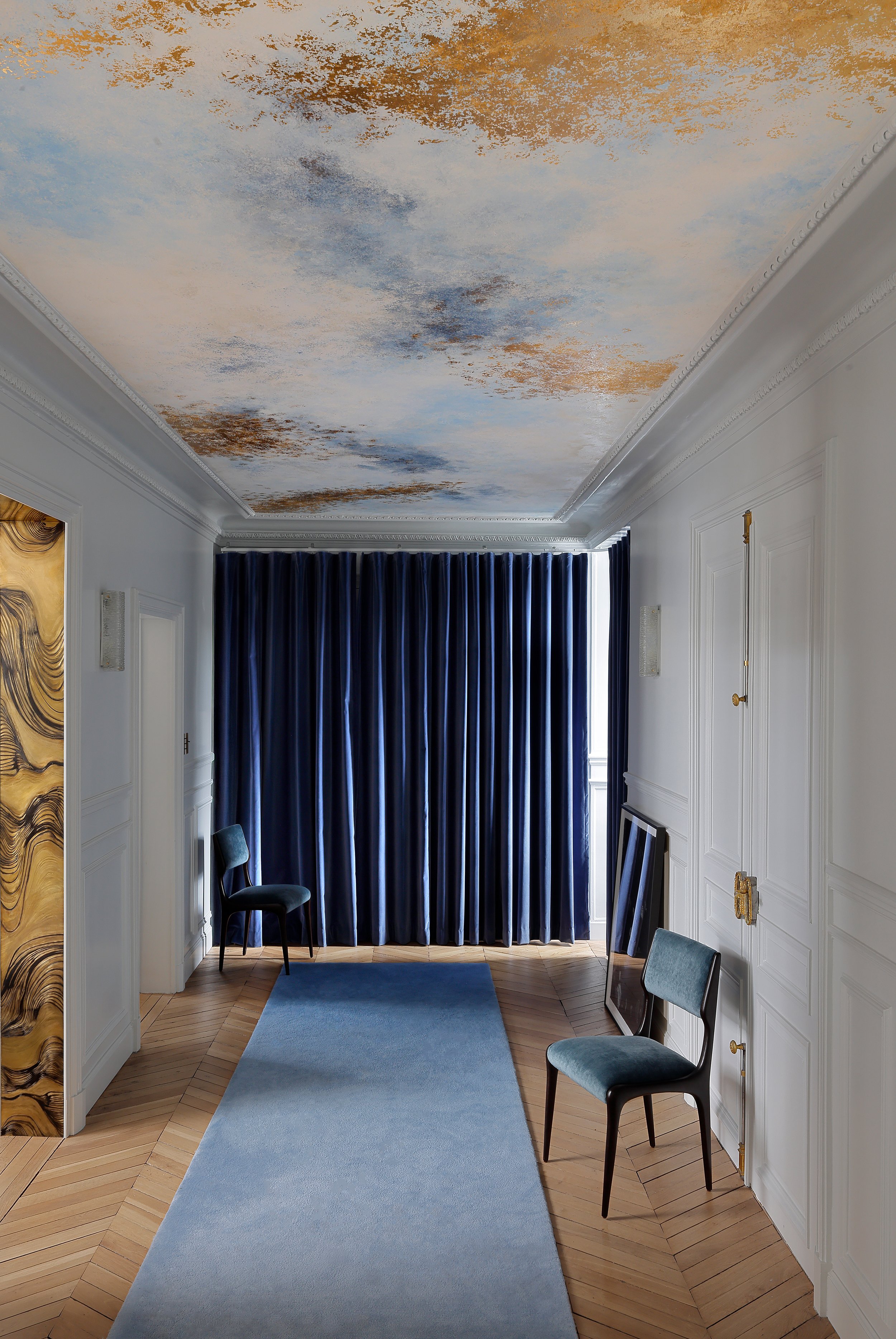 Une pièce élégante avec un plafond peint artistiquement évoquant un ciel nuageux, des rideaux en velours bleu foncé couvrant une fenêtre, un tapis bleu clair au sol, des chaises en tissu bleu clair, un tableau posé contre le mur, et des murs blancs avec moulures décoratives.