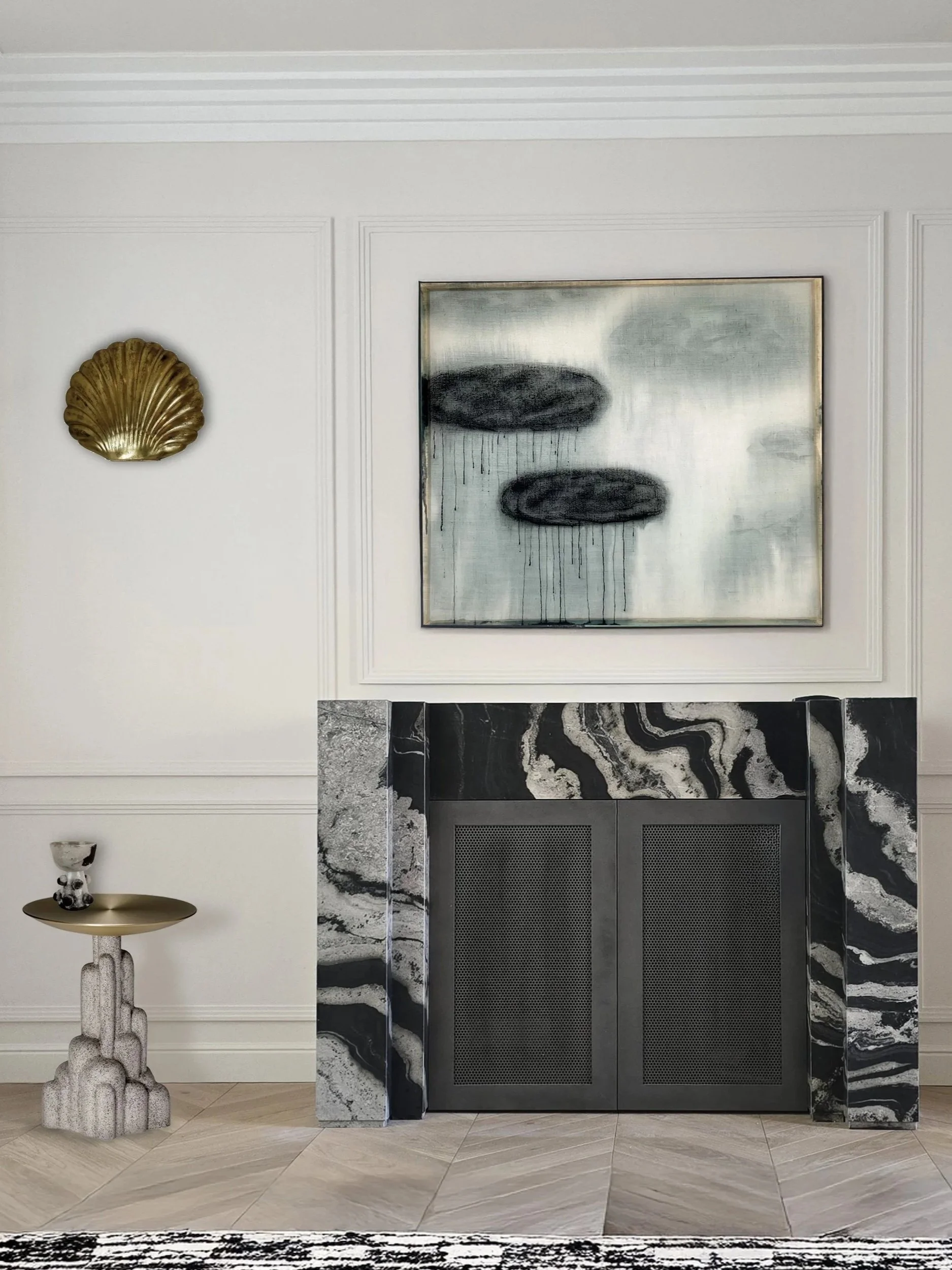 A white wall with a black-and-gray abstract painting above a black-and-white marble fireplace. To the left, a small stone table with a ceramic object, a black fireplace and a gold shell decoration.