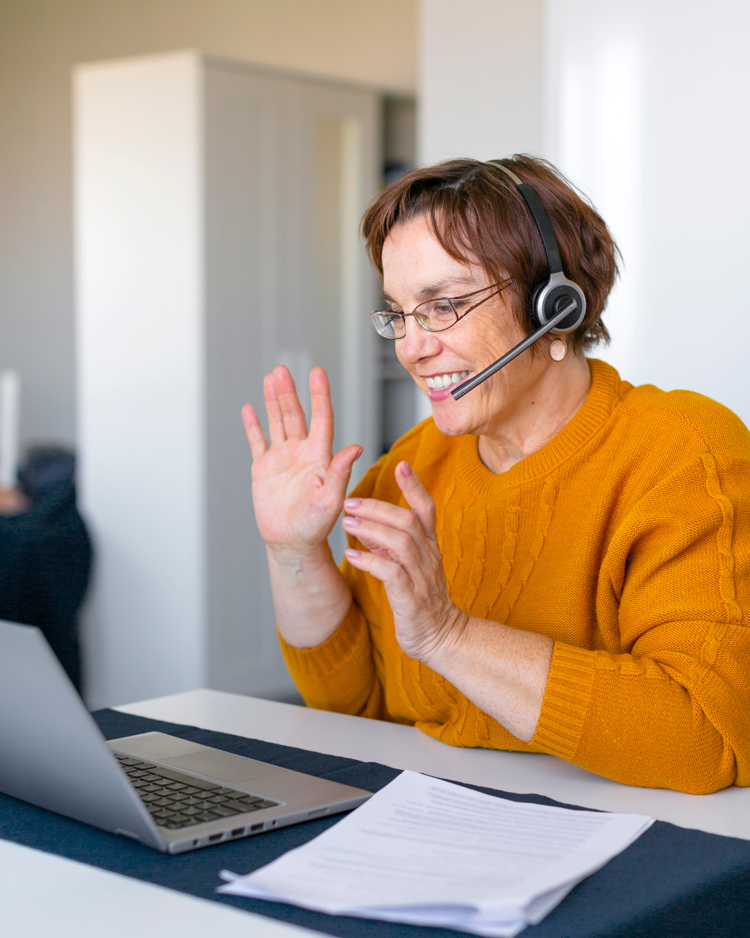 A female business owner with short brown hair, glasses, and wearing a yellow sweater, is smiling and waving while joining in an online community event. She is wearing a headset with a microphone.