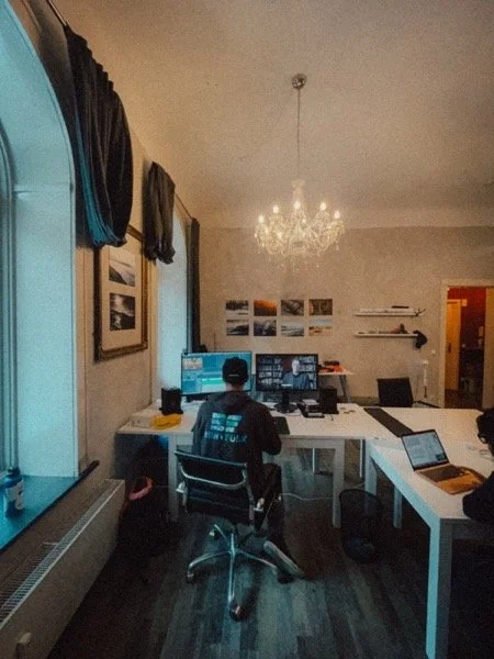 Jemand arbeitet an einem großen Schreibtisch im Arbeitszimmer, mit mehreren Computern und einem Laptop, beleuchtet von einem Kristallchandelier, Wände mit Fotos und Regalen, dunkle Vorhänge an den Fenstern.