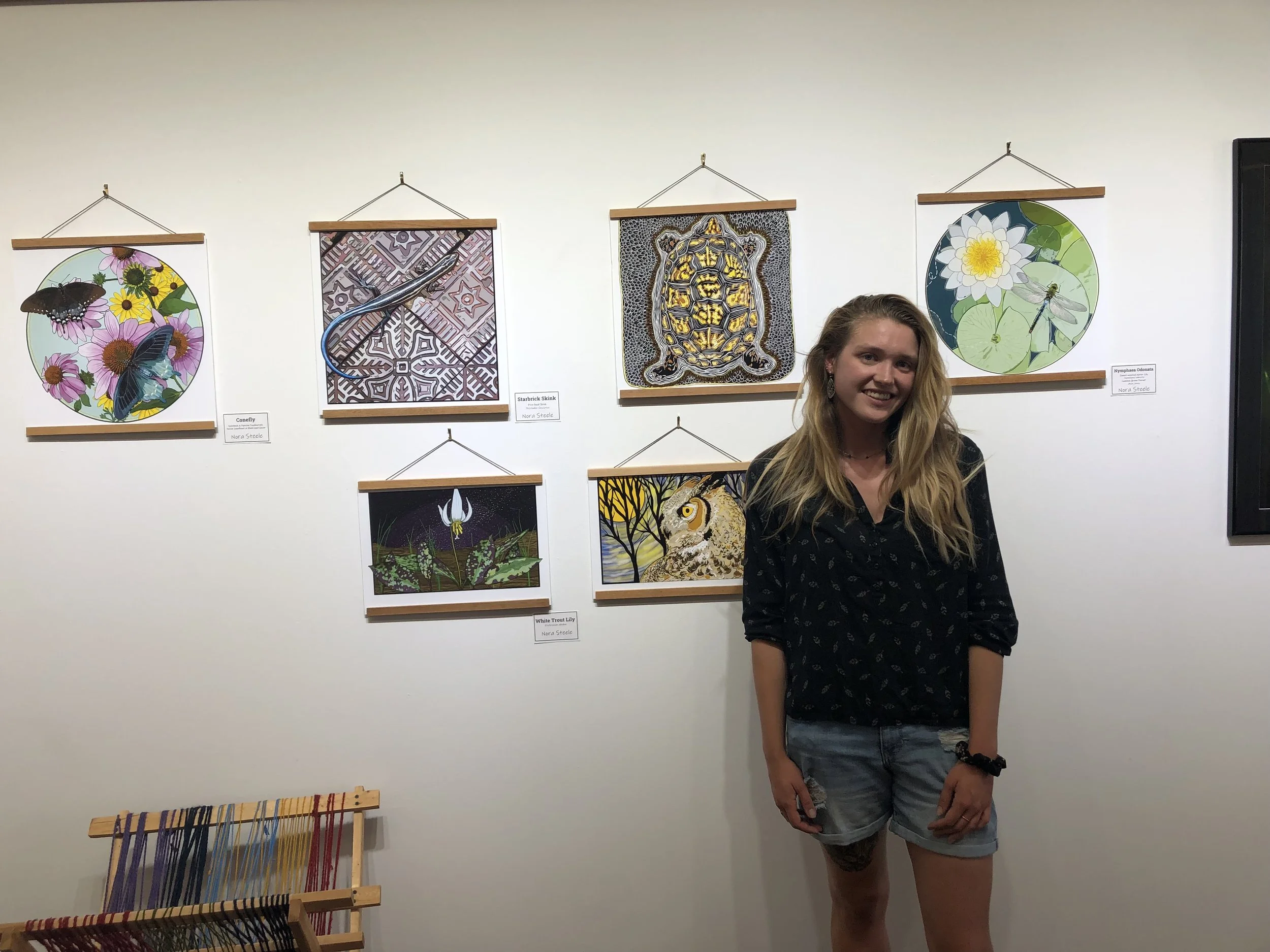 A young woman with long blonde hair, wearing a black shirt with a pattern and denim shorts, stands smiling in front of a wall displaying colorful artwork and photographs of insects and animals, with a small woven art display at the bottom left.