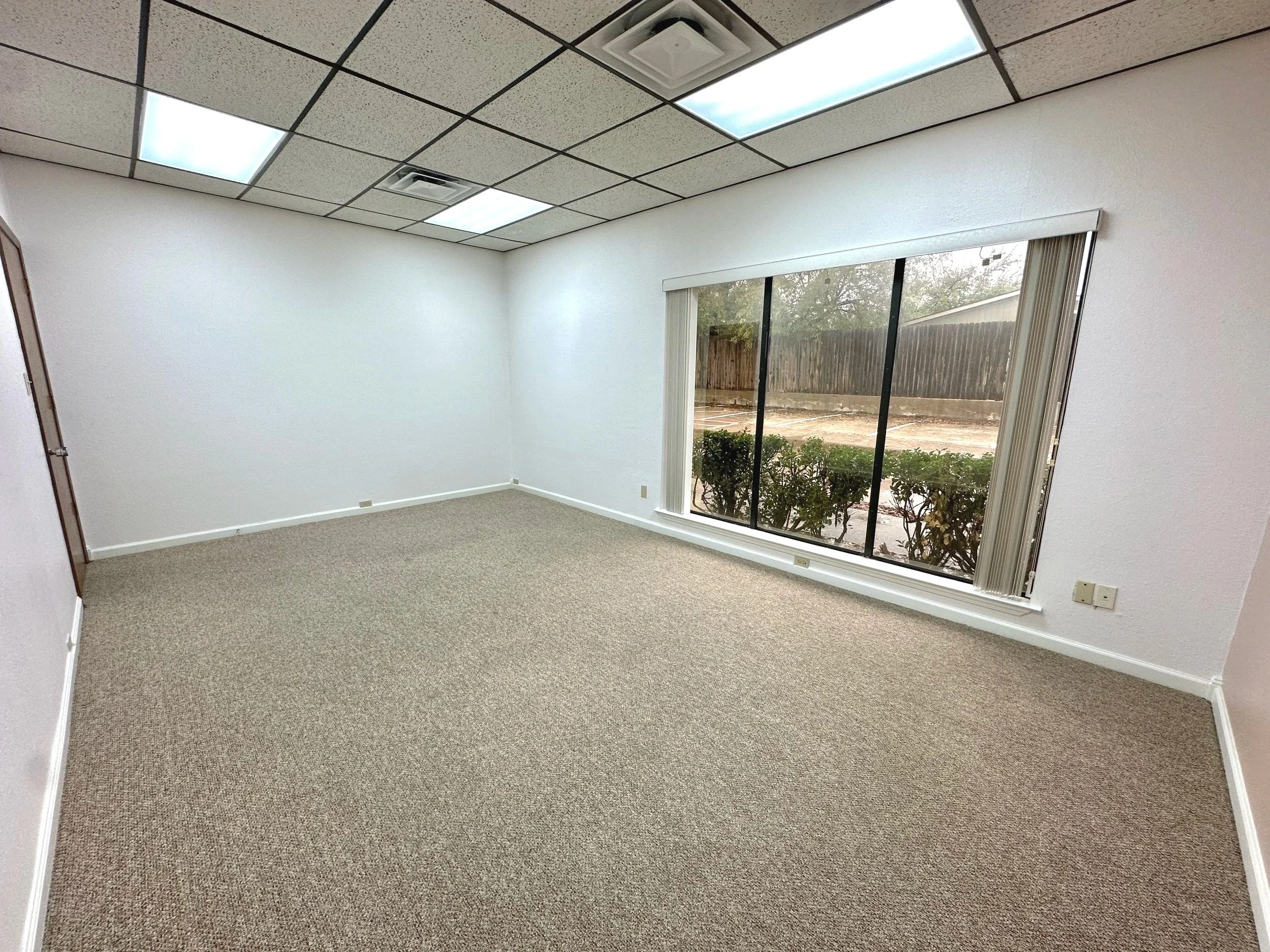 Empty office room with beige carpet, white walls, large window with blinds, and a drop ceiling with fluorescent lights.