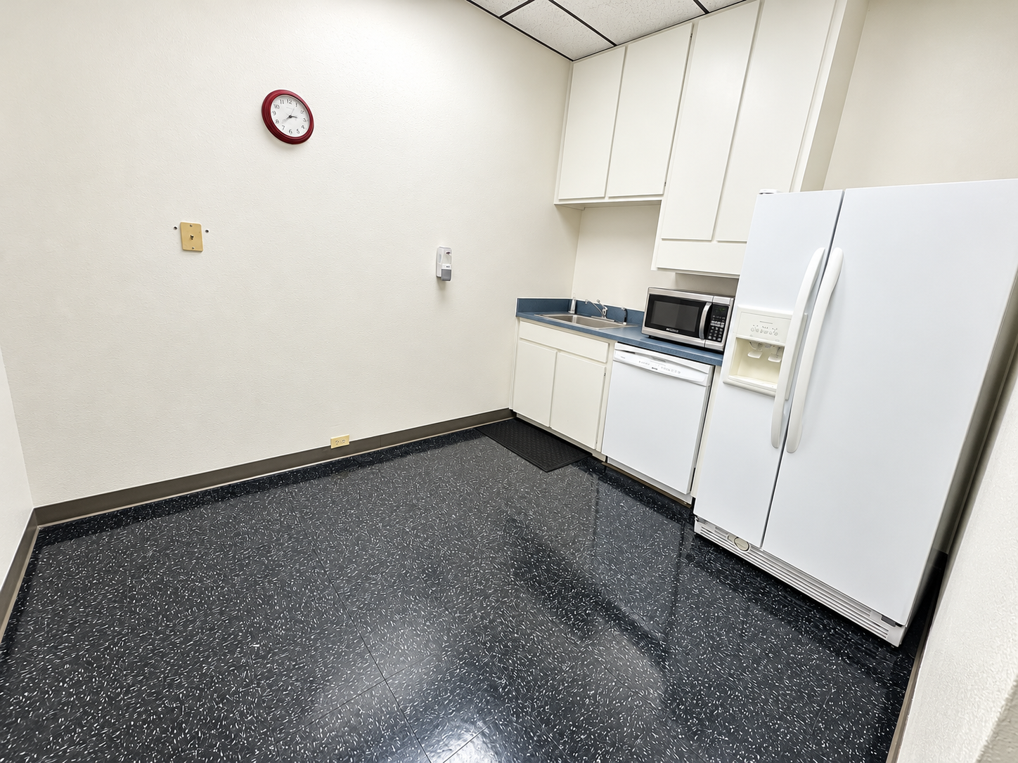 Office kitchenette with white cabinets, refrigerator, and dark flooring at 6825 Manhattan Boulevard in Fort Worth, Texas.