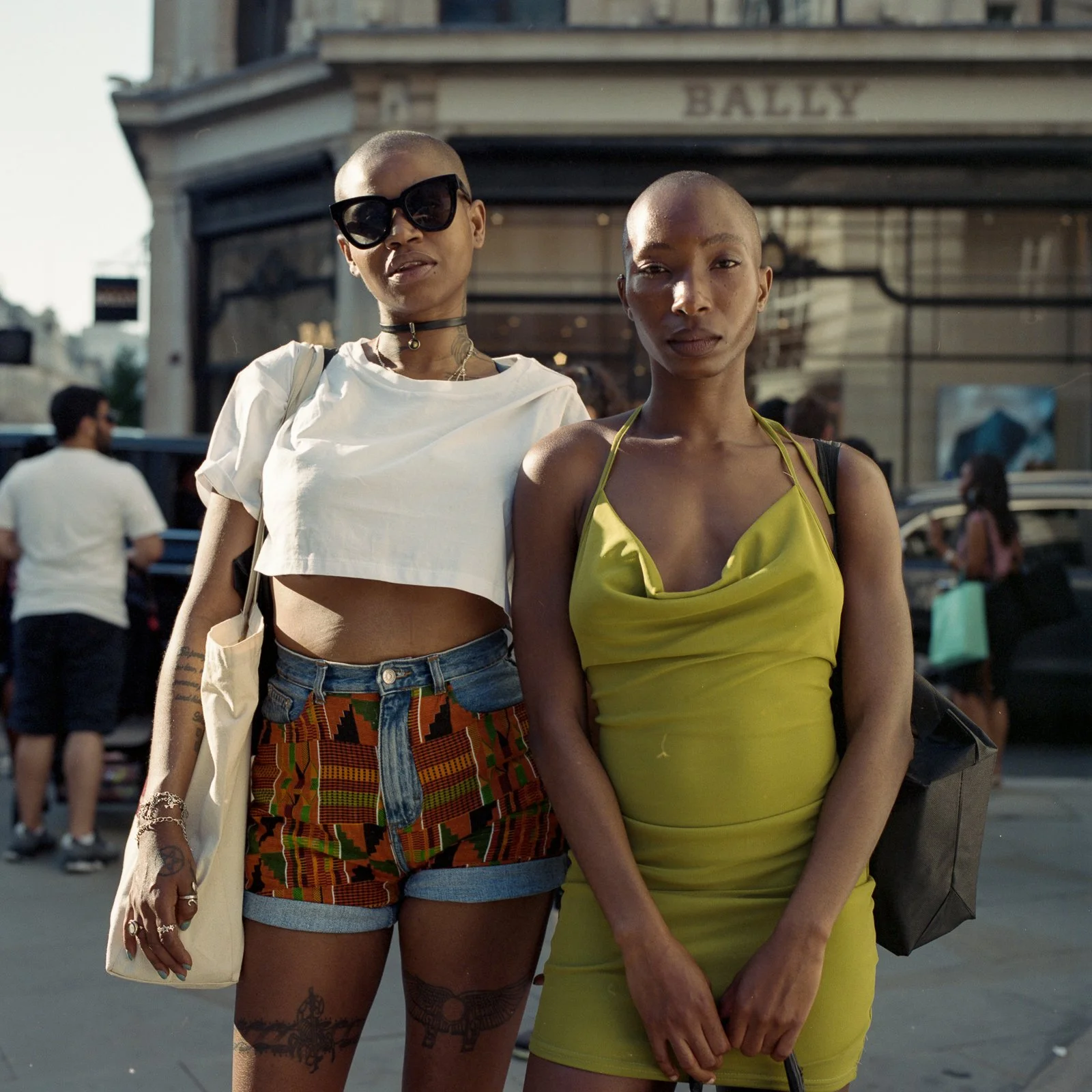 Two women standing together outdoors, one wearing sunglasses, a crop top, and patterned shorts, and the other wearing a green dress, with a city street and storefront in the background.