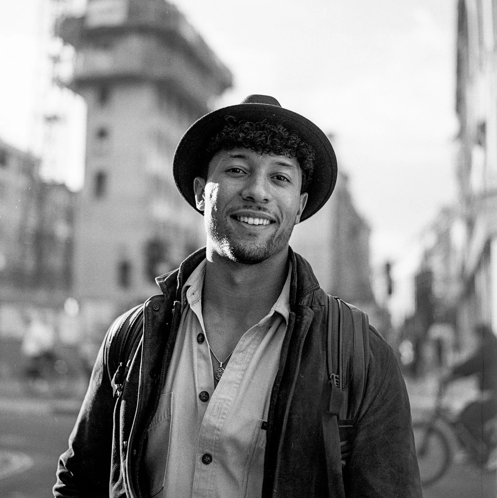 A young man with curly hair, wearing a hat, smiling, outdoors in an urban setting, in black and white.