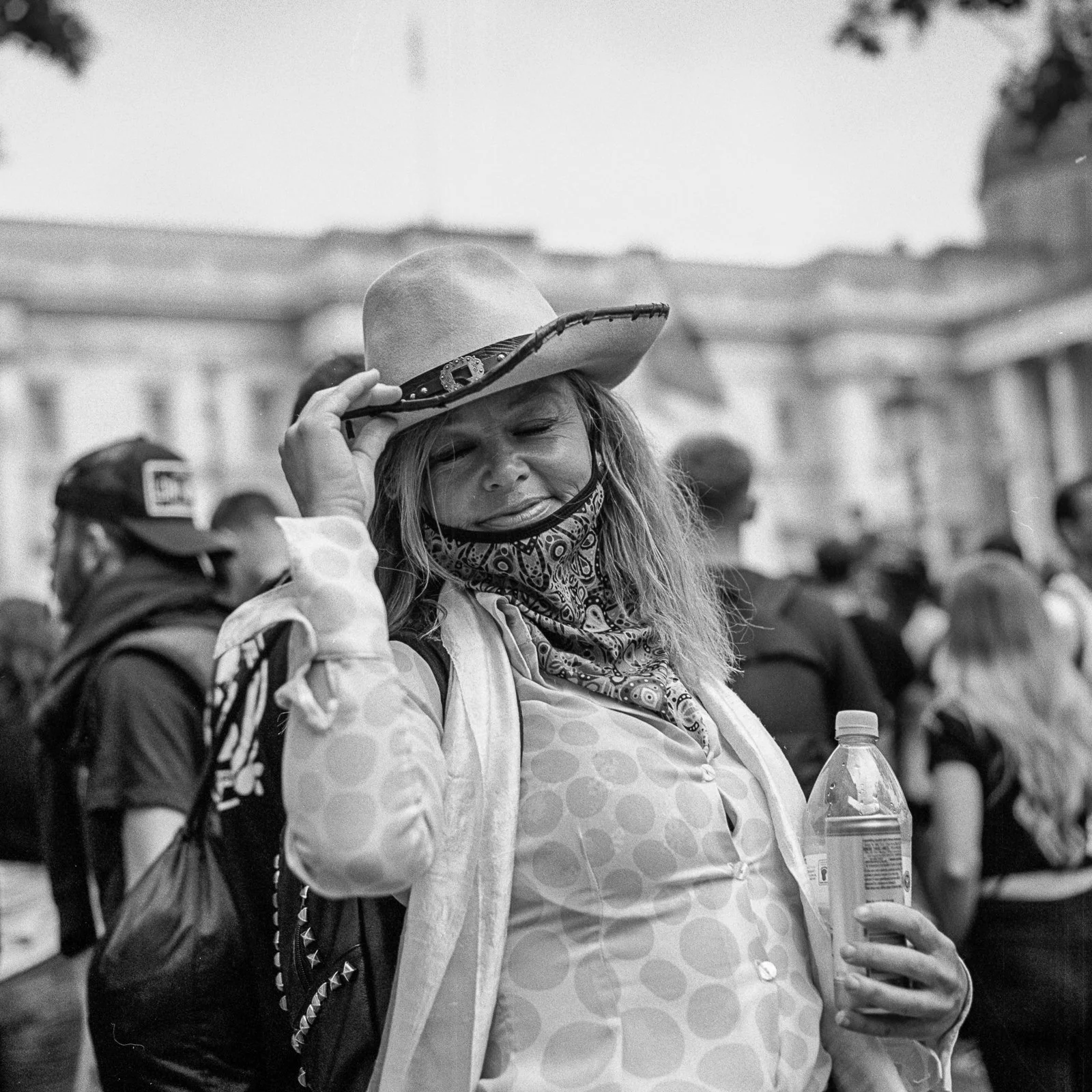 A woman wearing a cowboy hat and a bandana around her neck, holding a water bottle and adjusting her hat in a crowd outdoors.