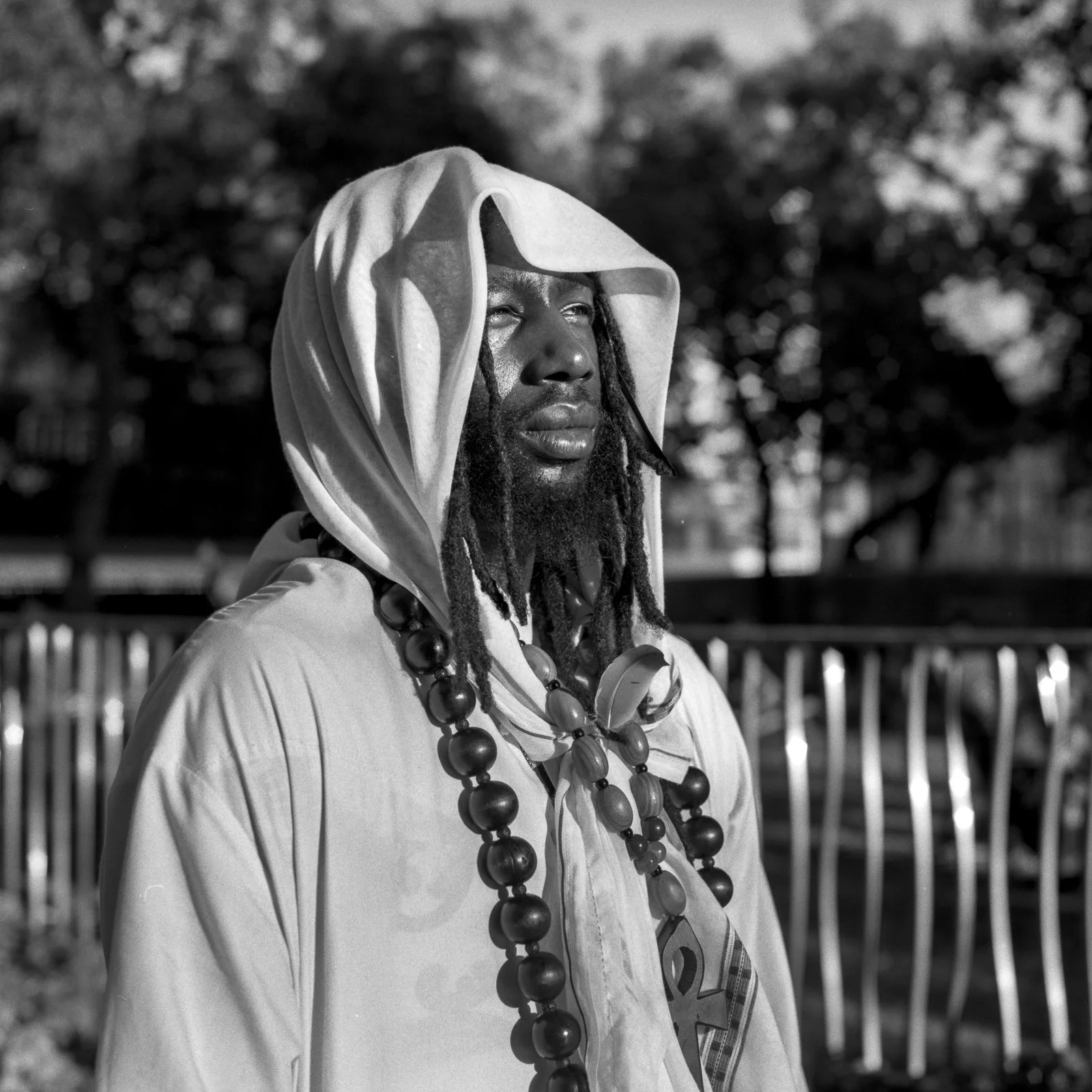 A man with a hood draped over his head, wearing necklaces, standing outdoors with trees and a fence in the background.