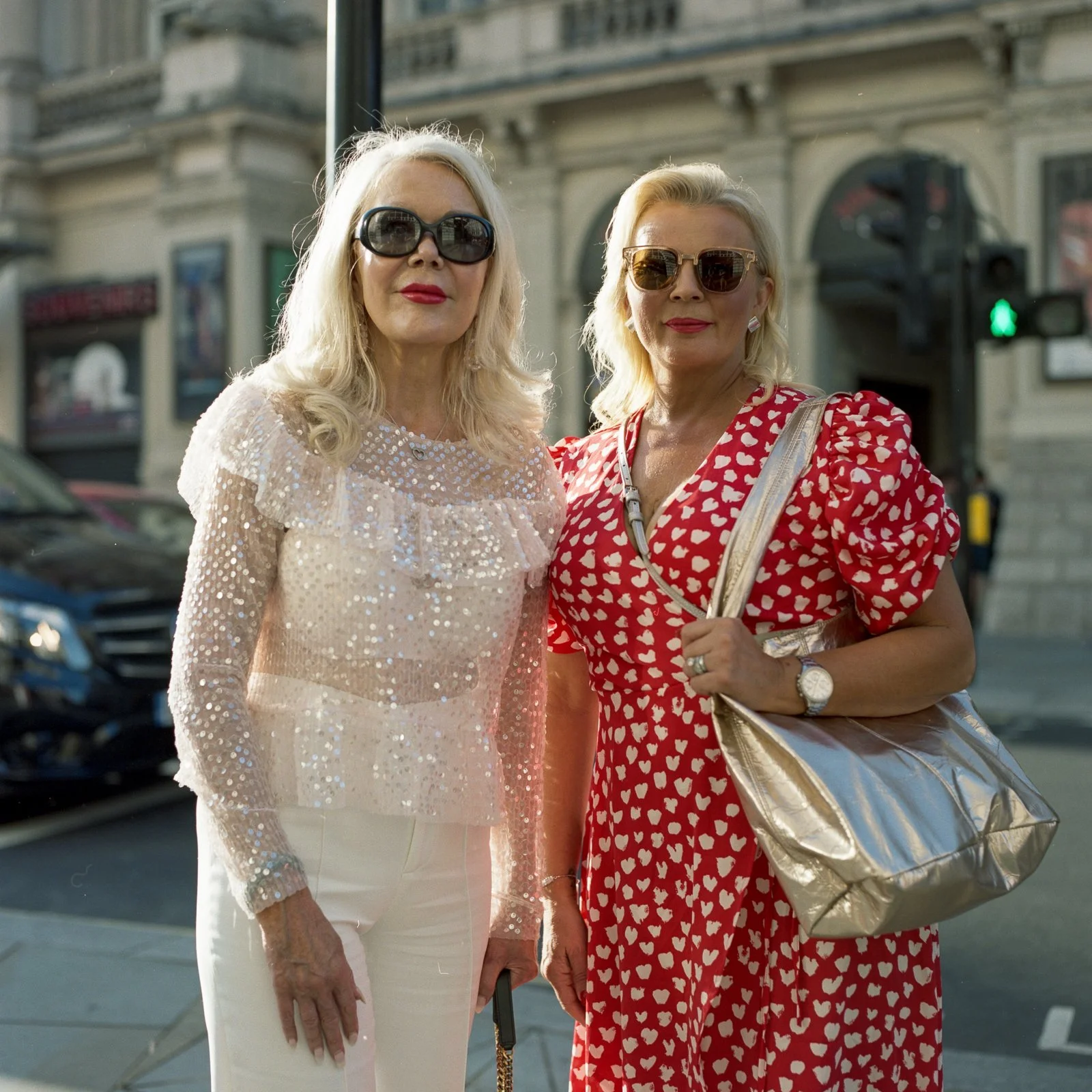 Two women standing on city street, wearing fashionable outfits and sunglasses, with a city building and traffic light in background.