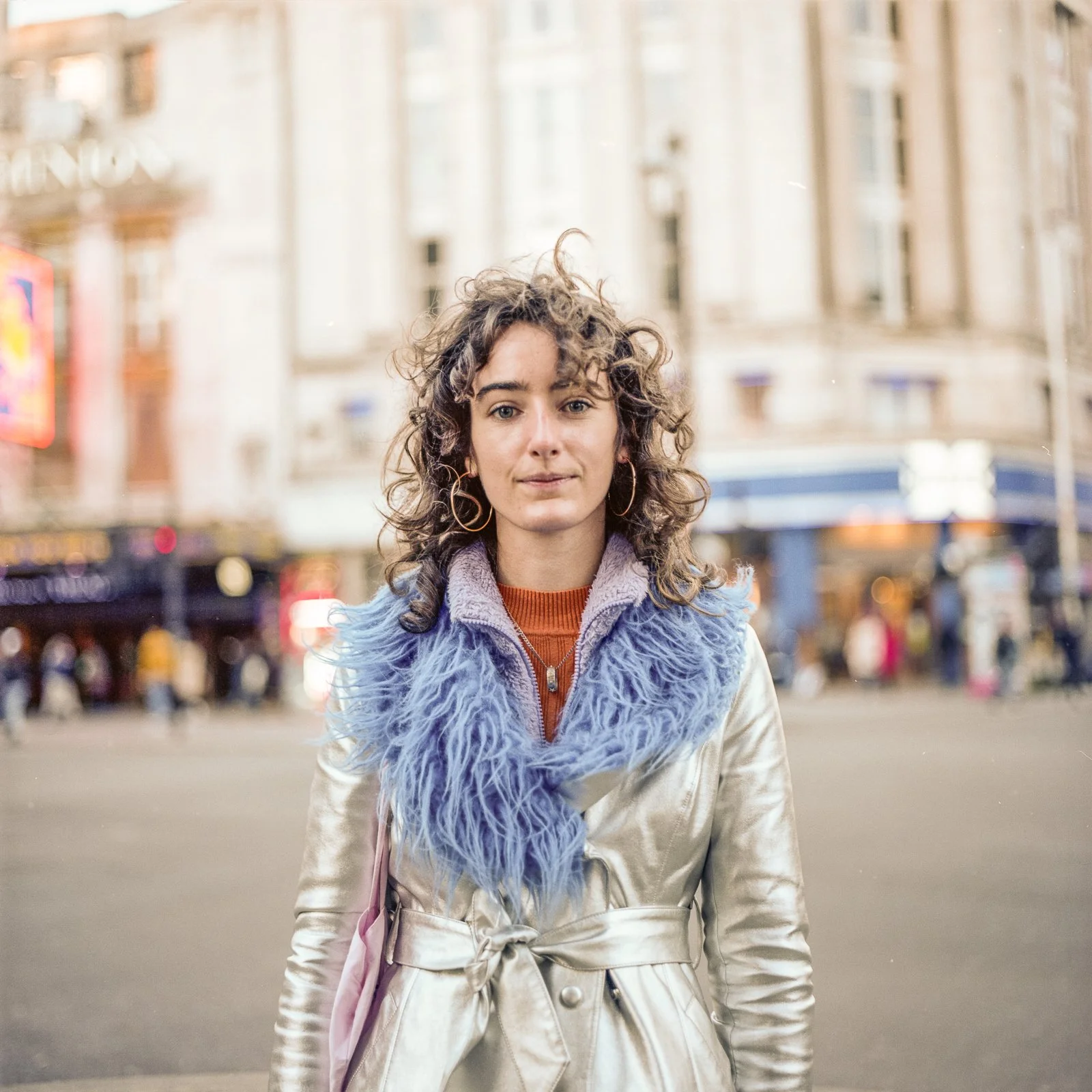 A woman standing on a city street in front of a busy background with buildings and pedestrians, wearing a shiny beige coat with a blue faux fur collar and hoop earrings.