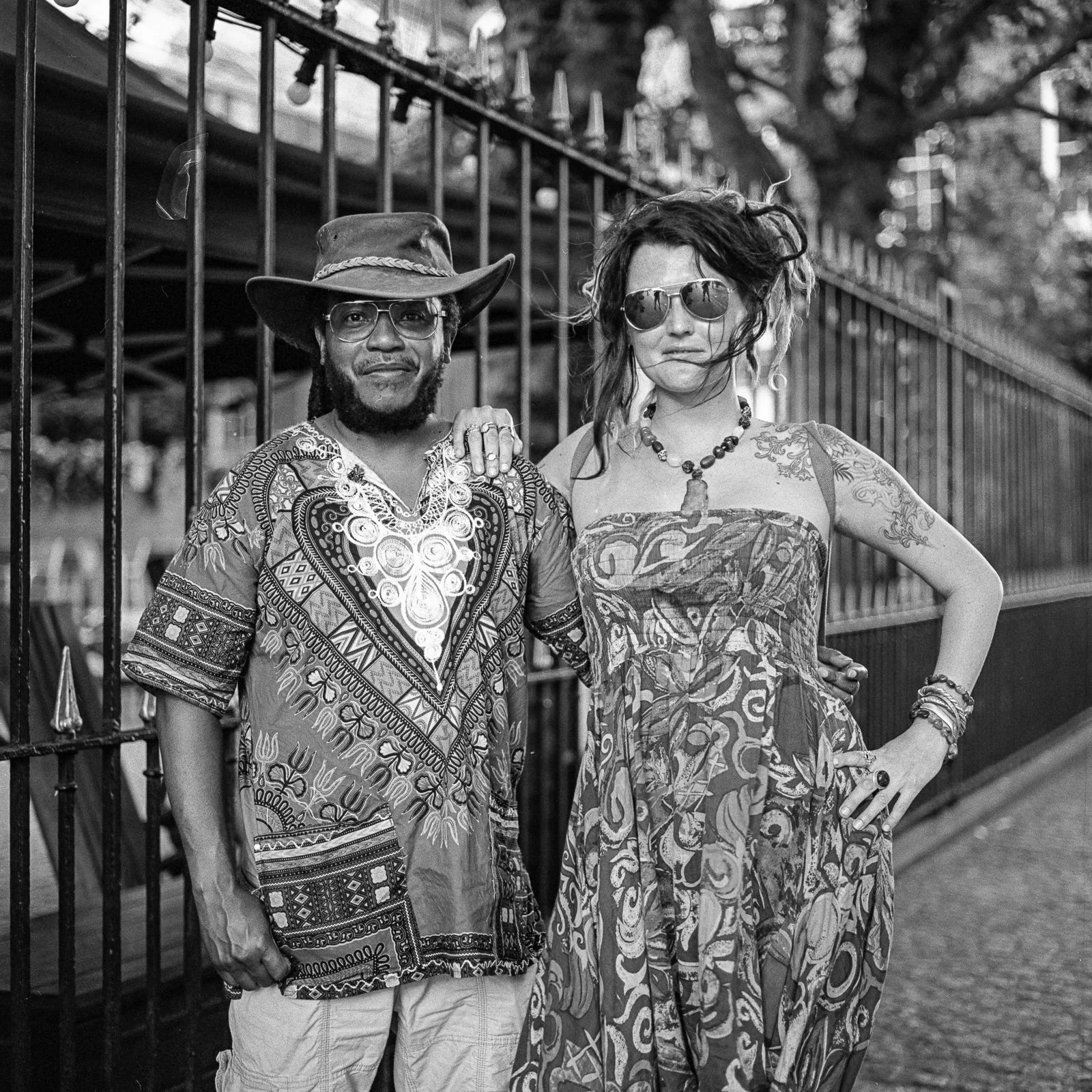 Two people standing outdoors in front of a metal fence. The person on the left is a man wearing glasses, a wide-brimmed hat, and a patterned shirt. The person on the right is a woman with wavy hair, wearing sunglasses, a strapless dress, and various jewelry, with her hand on her hip.