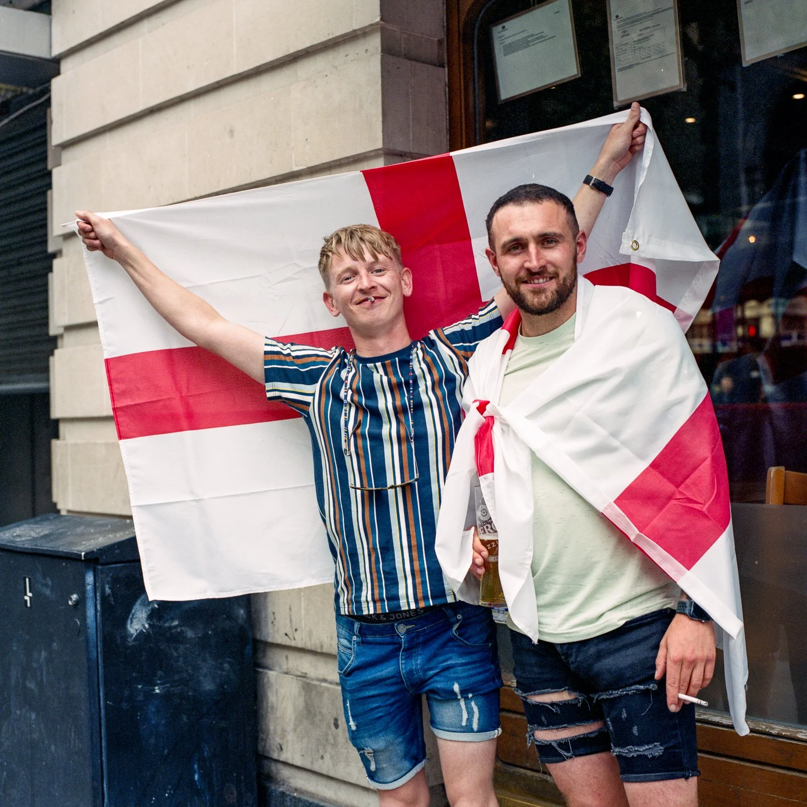 Two men standing in front of a building, celebrating with an English flag draped around one of them. One man is smiling with his arm outstretched holding the flag, and the other man is smiling, holding a drink, with the flag draped over his shoulders.