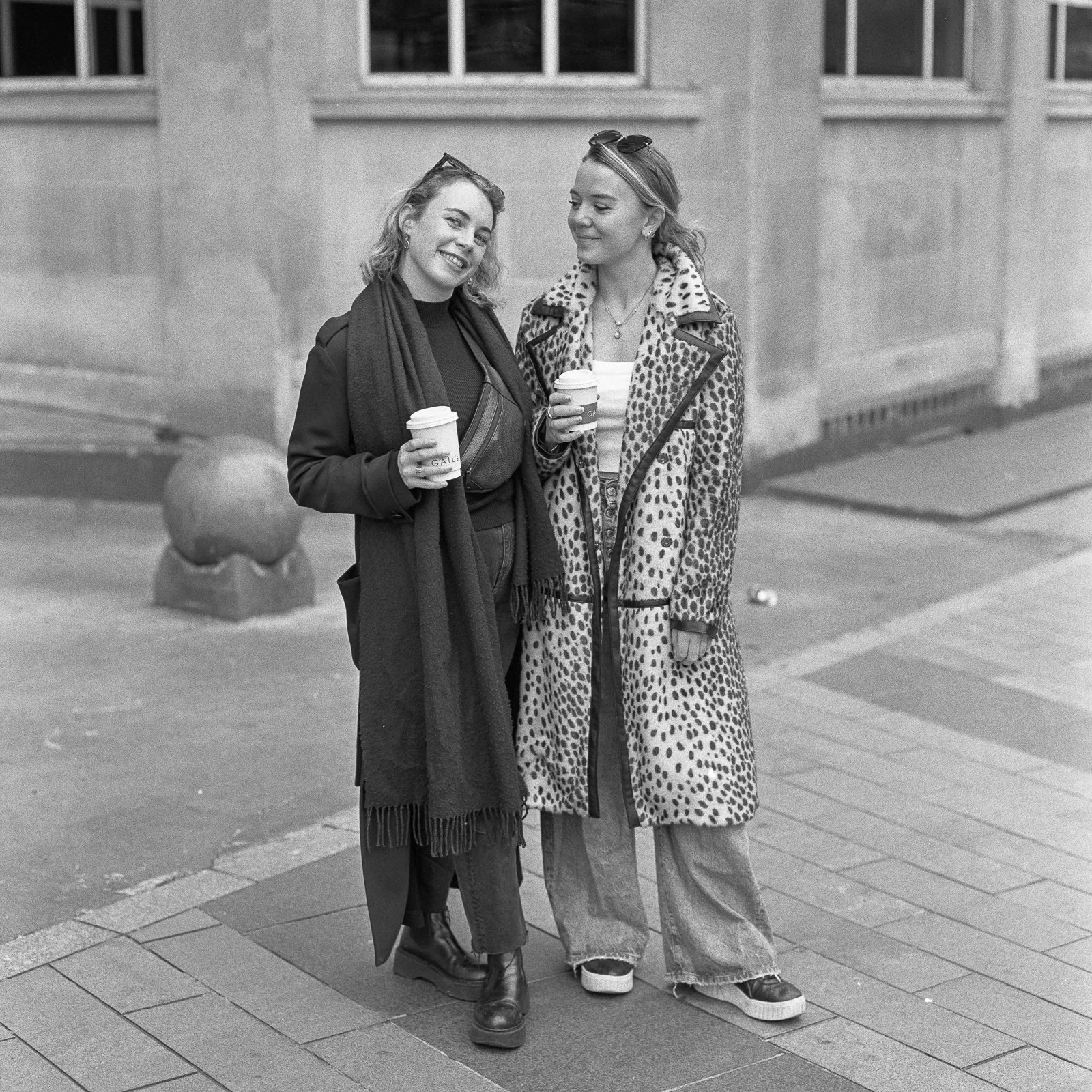 Two women standing outdoors holding coffee cups, smiling, and looking at each other, with a stone building in the background.