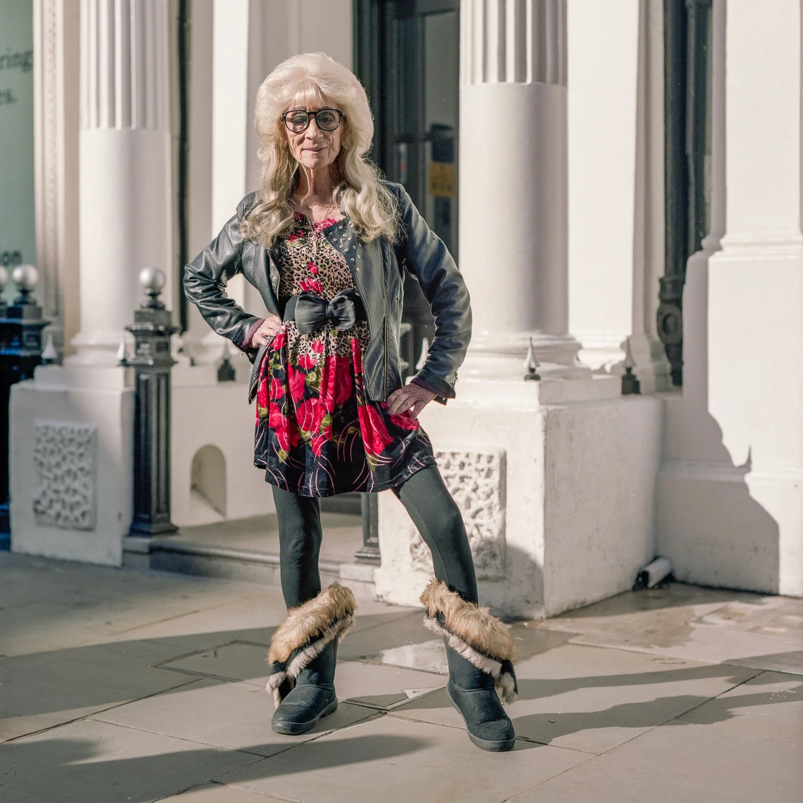 An older woman wearing a black leather jacket, colorful dress, black tights, and furry boots stands with her hands on her hips in front of a white building with tall columns.