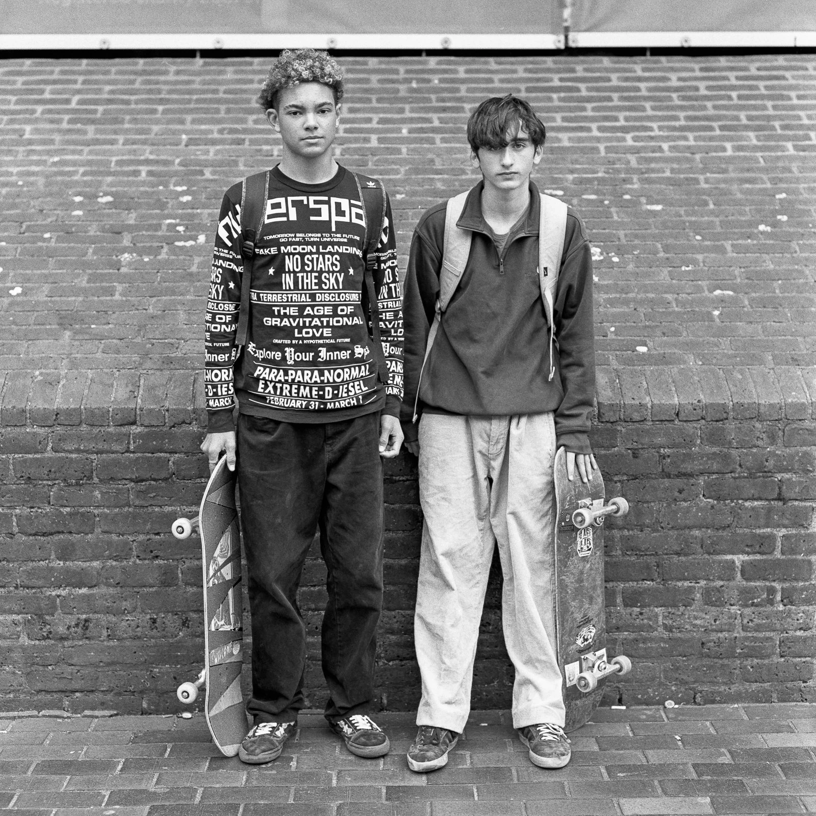 Two teenage boys standing against a brick wall holding skateboards.
