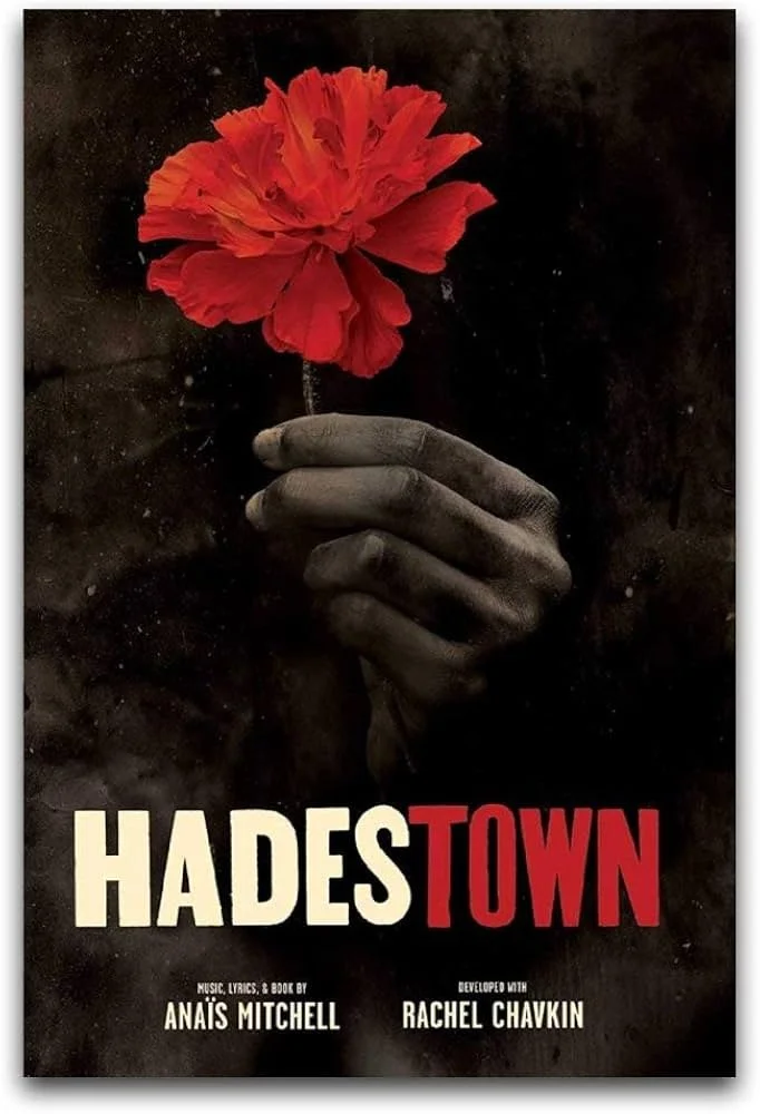 A dark poster featuring a hand holding a red carnation flower. The title 'Hades Town' is in large letters, with 'Hades' in white and 'Town' in red. The poster also credits Anais Mitchell for music, lyrics, and book, and Rachel Chavkin for development.