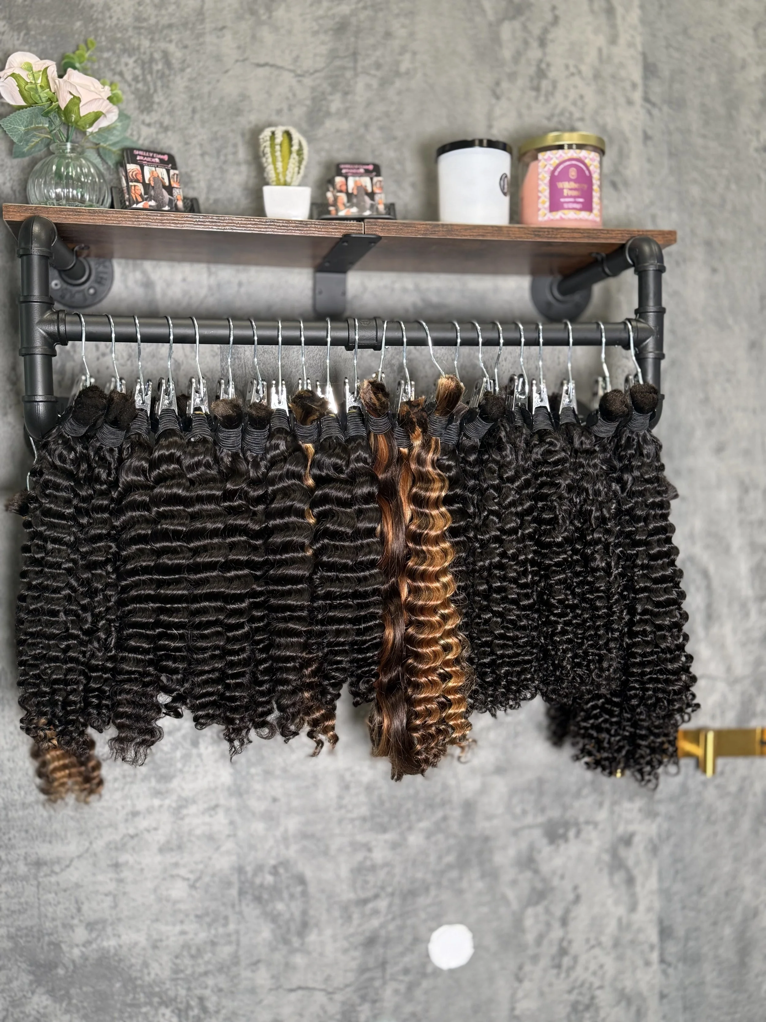 Multiple bundles of curly and wavy hair extensions hanging on a rack in a salon with a concrete wall background and a shelf with decorative items above.