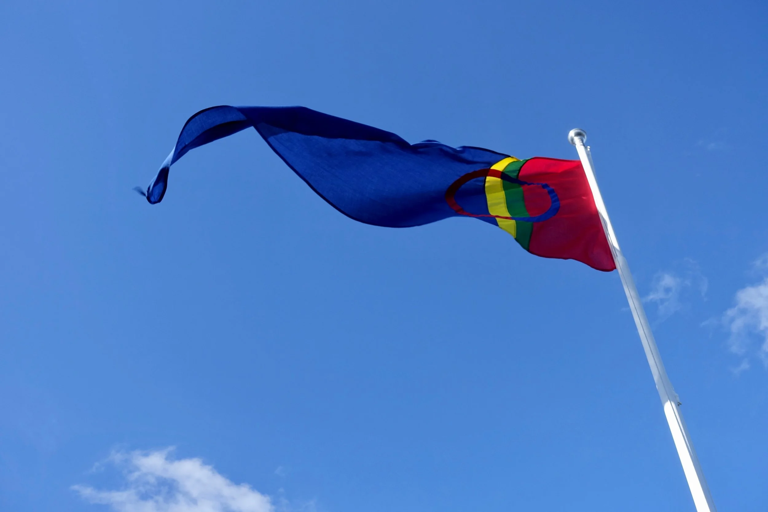A Sámi flag flying from a flagpole as seen from below, with a blue sky in the background.