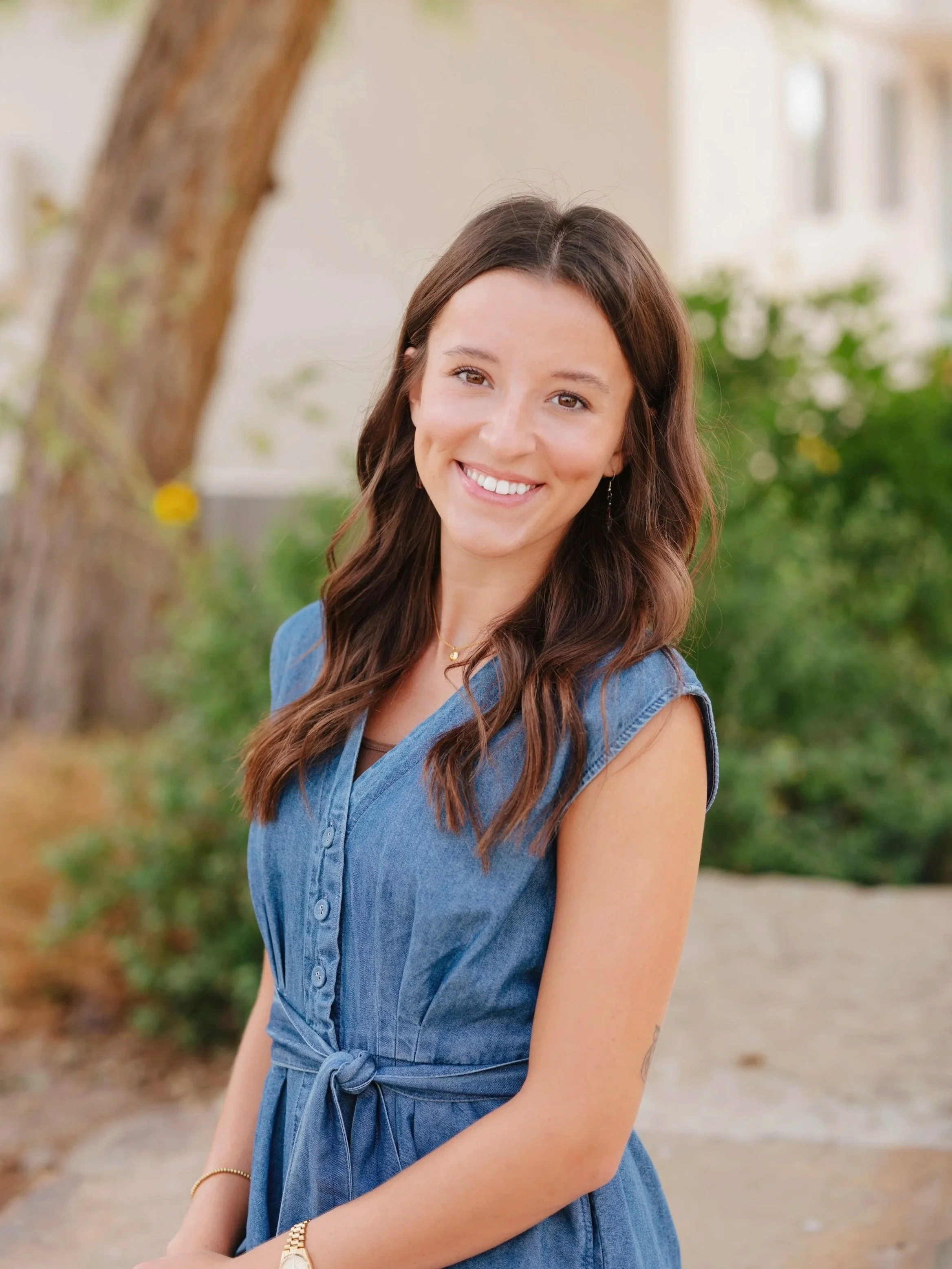 A young woman with long brown hair, smiling, wearing a denim dress with rolled sleeves, standing outdoors in front of a tree and blurred background of greenery and a building.