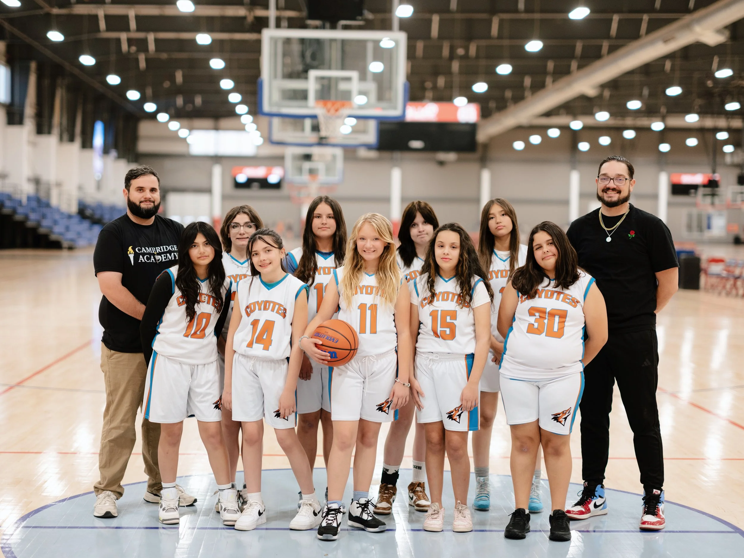 Young female basketball team with coaches on the court at an indoor sports arena. The players are wearing white uniforms with orange and blue accents, and the team name 'Coyote' is visible. One girl is holding a basketball. The coaches are standing on either side of the team, smiling.