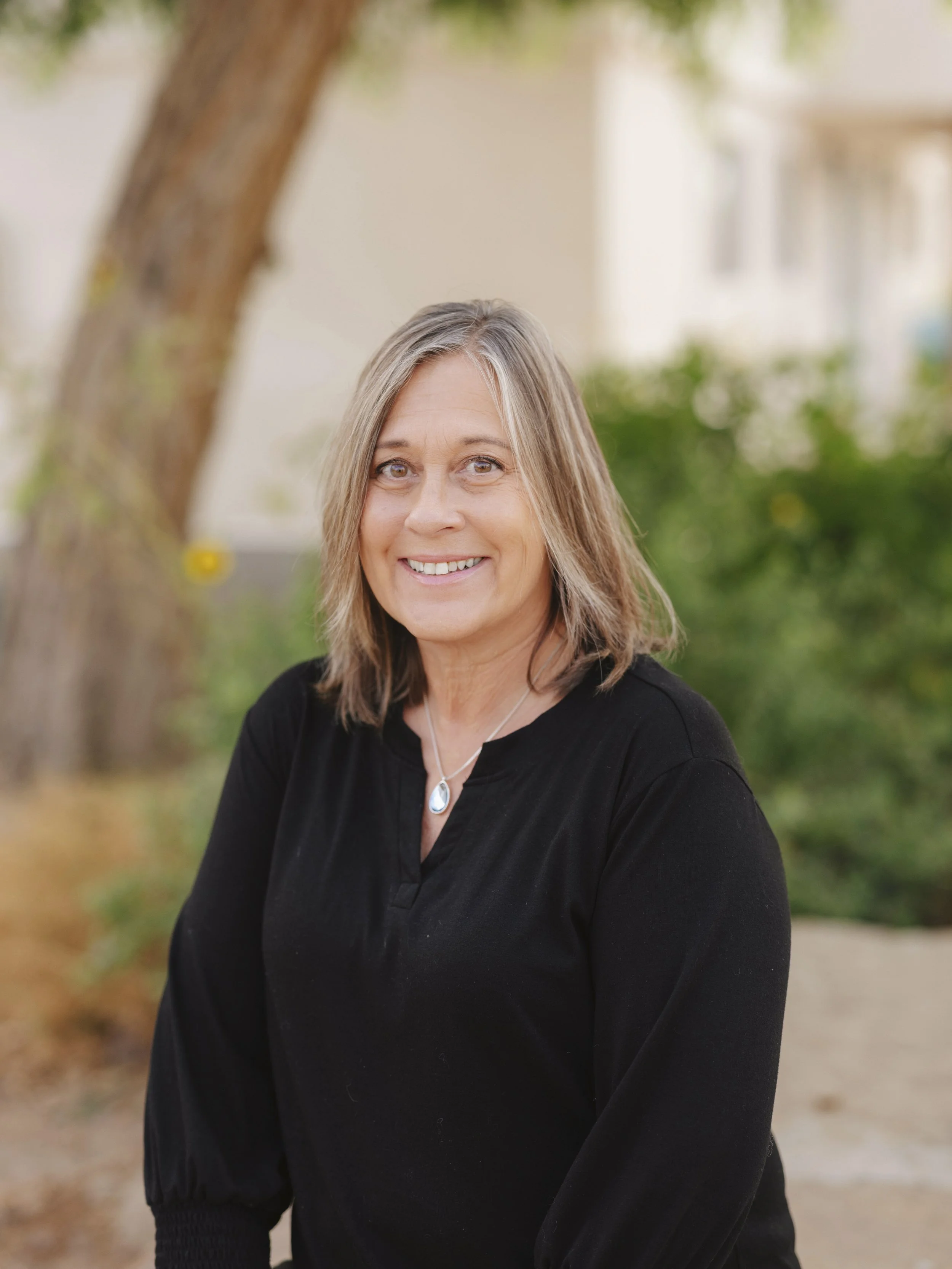 A middle-aged woman with shoulder-length gray hair, smiling, wearing a black top and a silver necklace with a teardrop-shaped pendant, outdoors with a blurred background of trees and greenery.