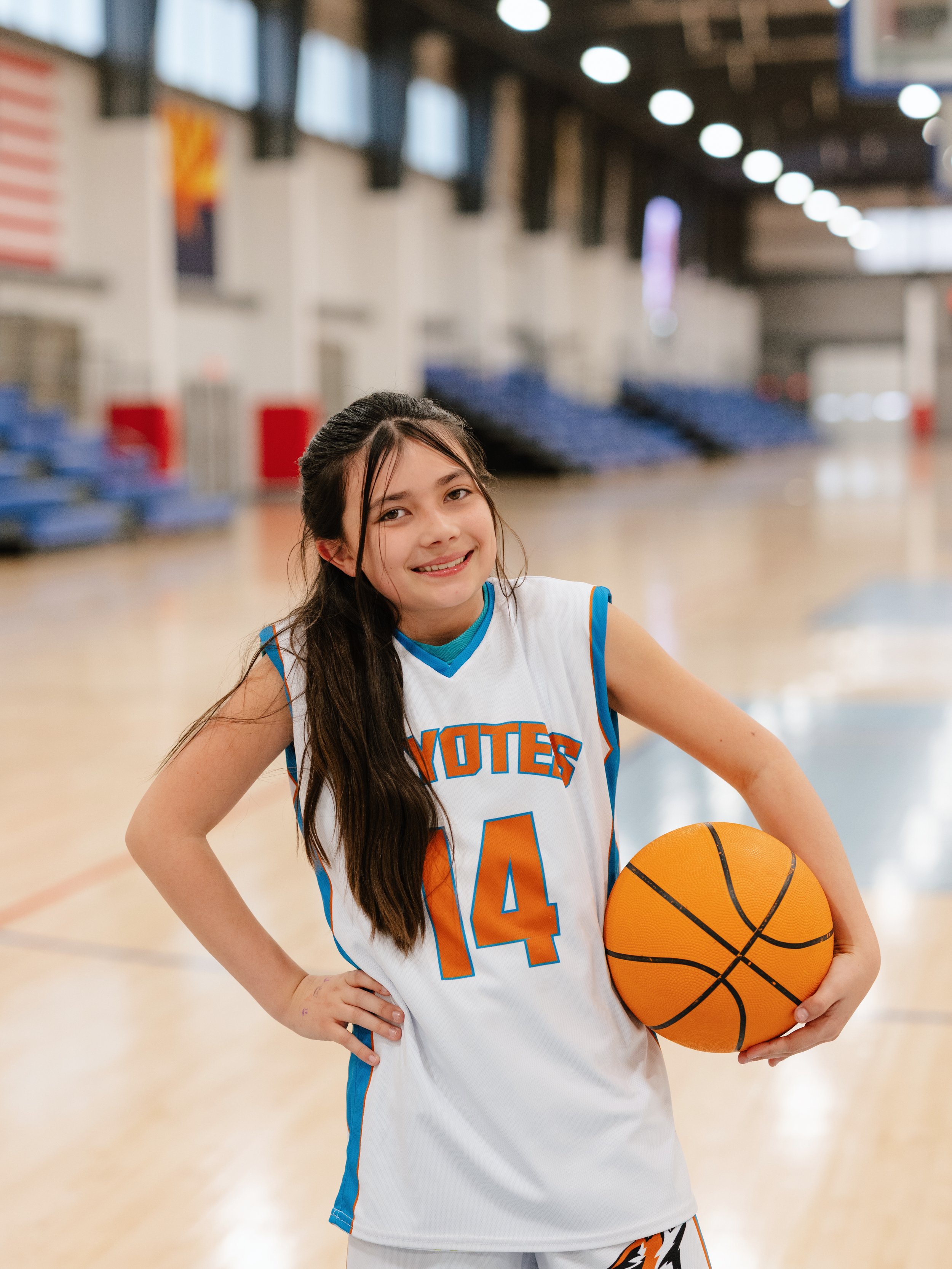 A young girl in a basketball jersey holding a basketball in a gymnasium.