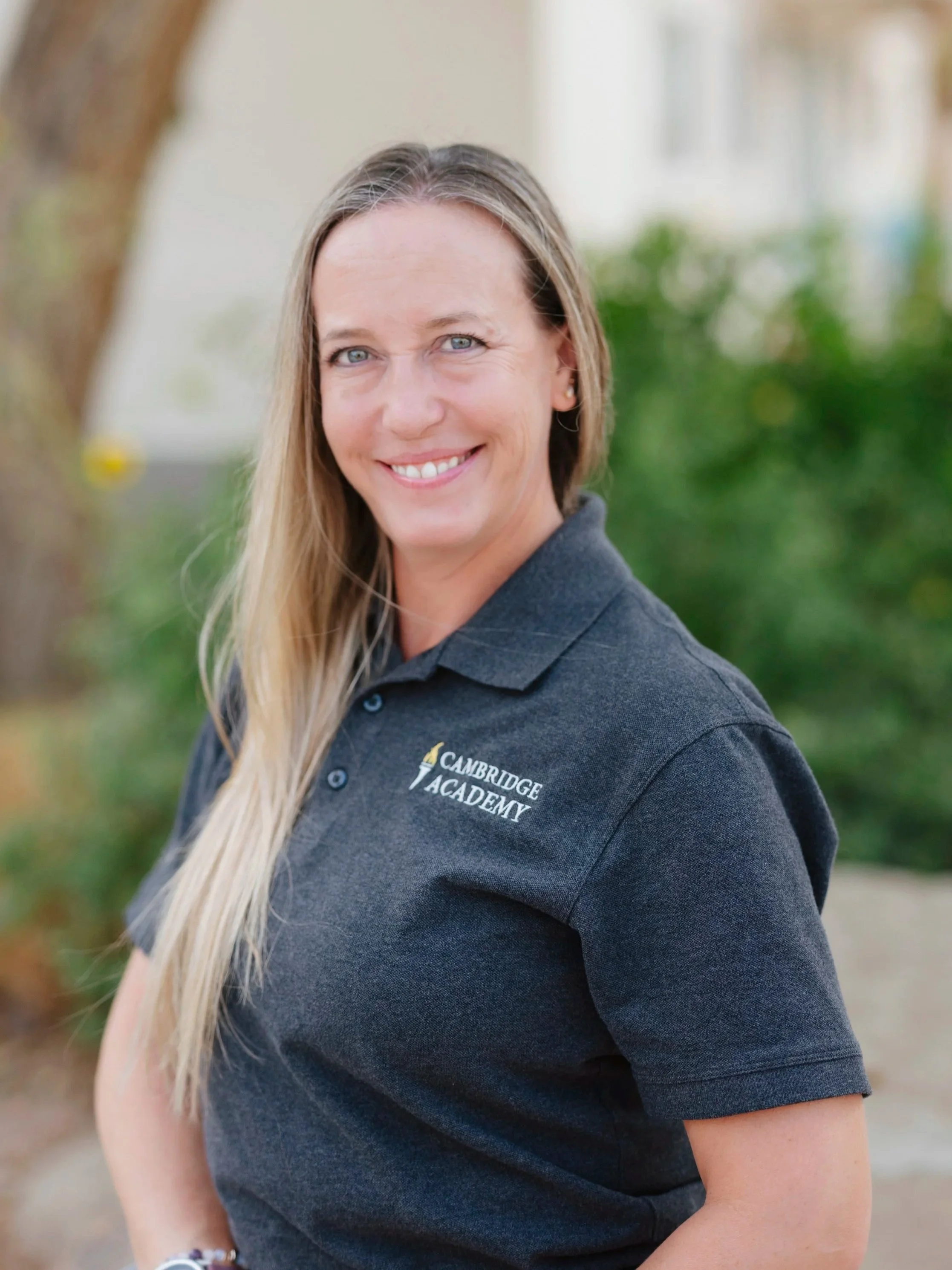 A woman with long blonde hair smiling outdoors, wearing a dark polo shirt with the logo 'Cambridge Academy' embroidered on it.
