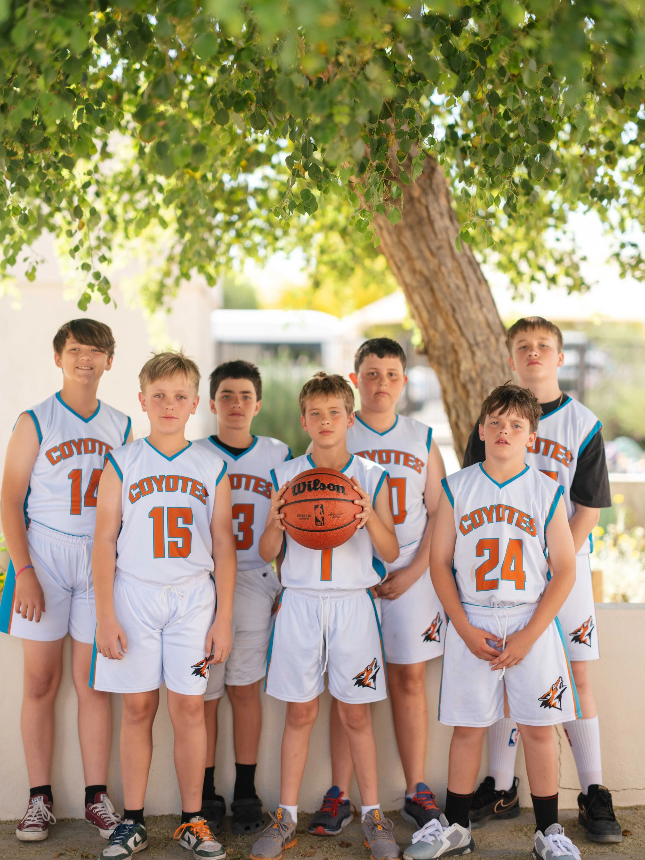 A group of seven young boys in basketball uniforms posing outdoors under a large tree. One boy in the center is holding a basketball, and they are all looking at the camera.