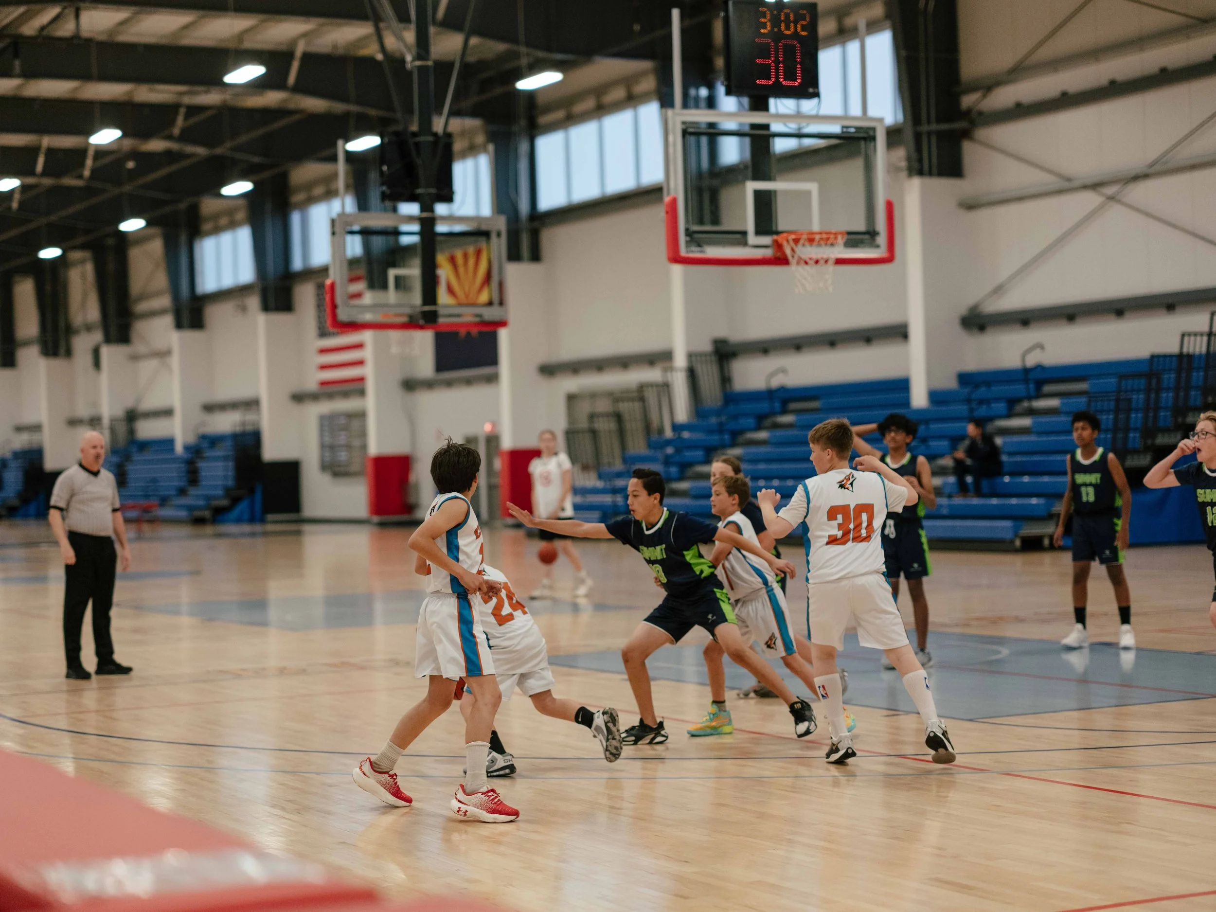 Youth basketball game in progress at indoor gym, players from two teams competing, referee nearby, scoreboard showing 3:02 on clock and 30 seconds, gym with blue bleachers and American flag in background.