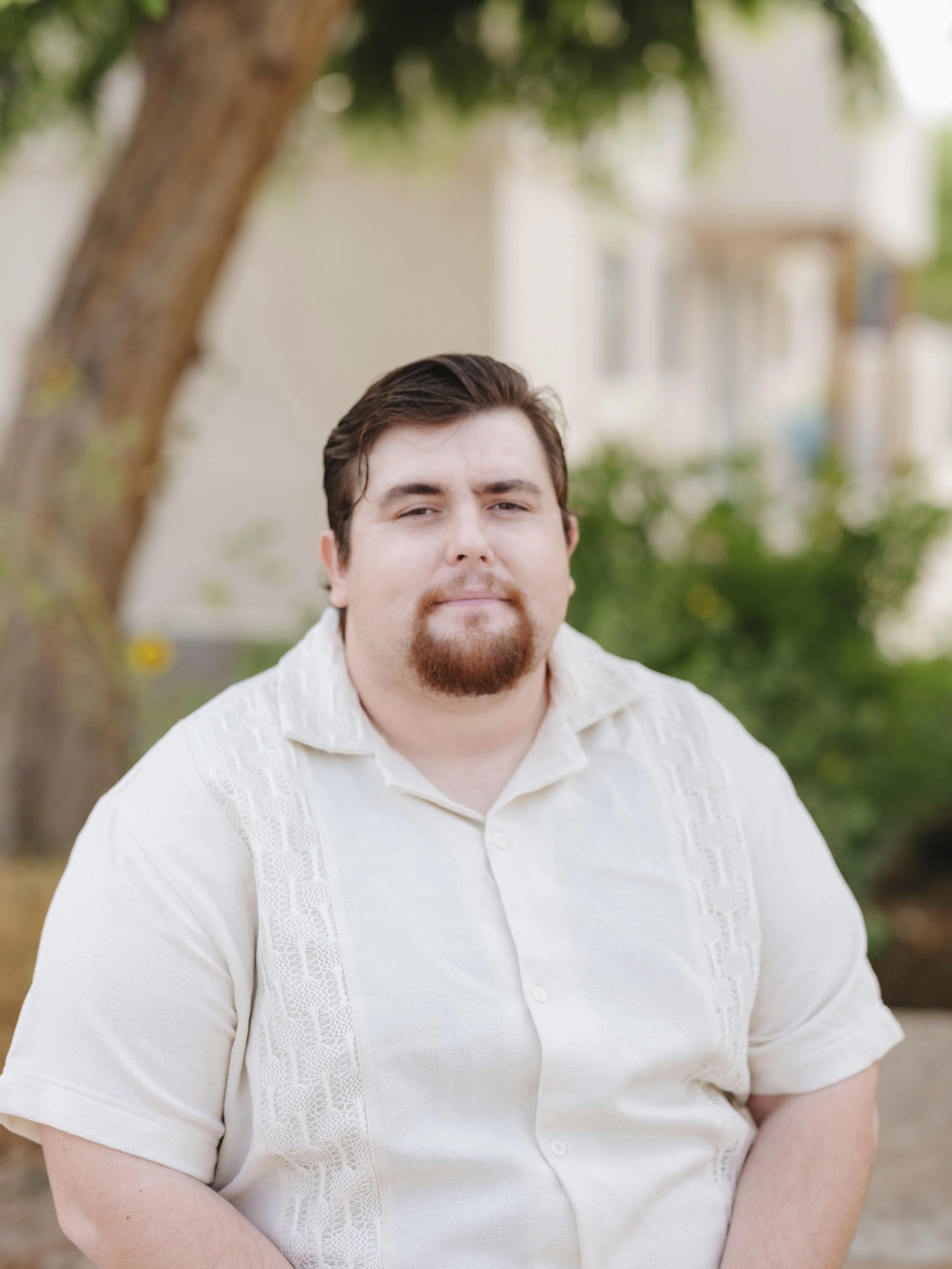 A man with dark brown hair and a beard, wearing a white shirt, standing outdoors near a tree with blurred buildings and greenery in the background.