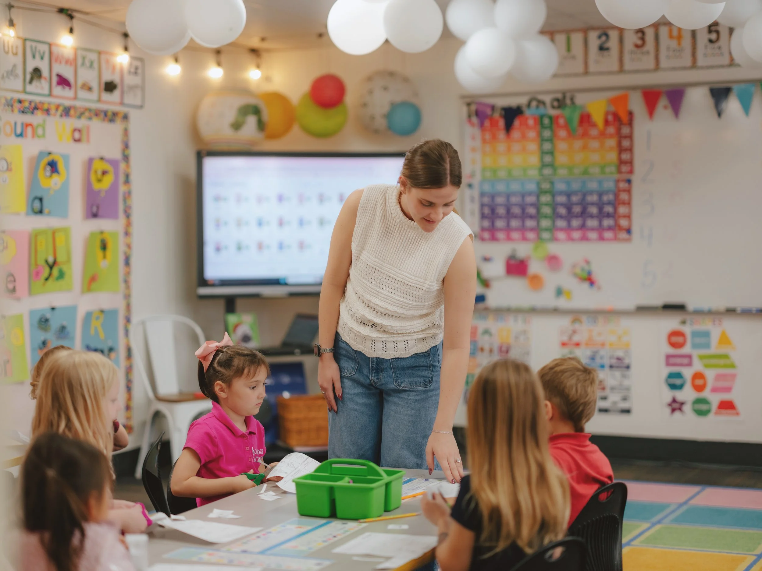 Classroom scene with a teacher and young students sitting at a table, colorful educational decorations on the walls, a large screen, and paper lanterns hanging from the ceiling.