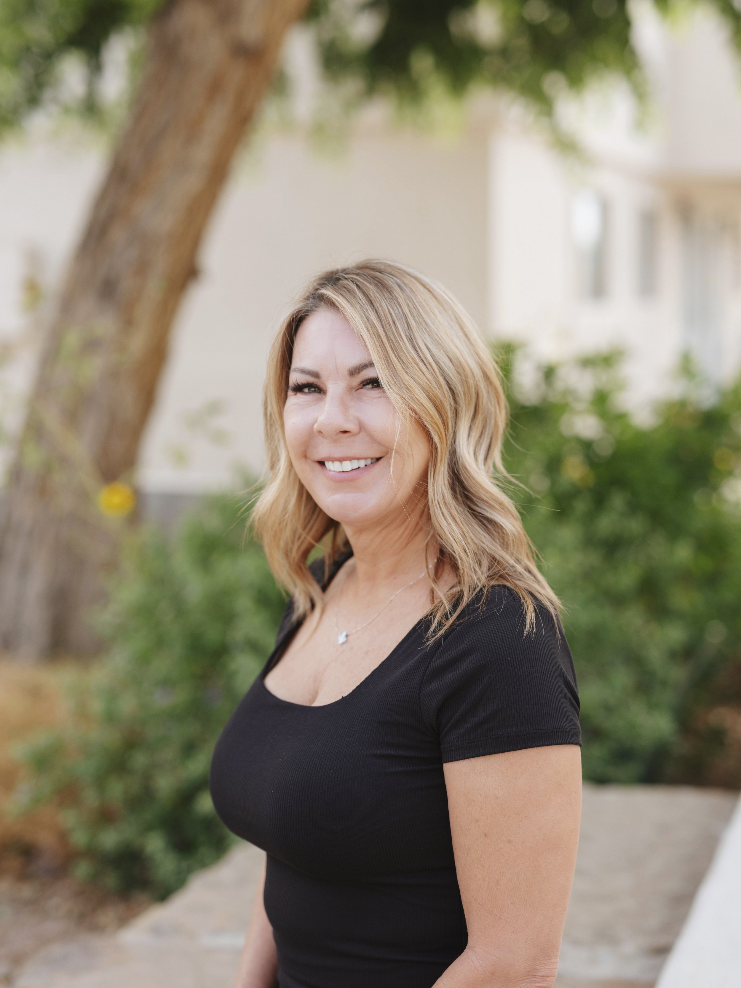 A smiling woman with shoulder-length blonde hair, wearing a black top, standing outdoors near a tree, with a blurred background of a white building and greenery.
