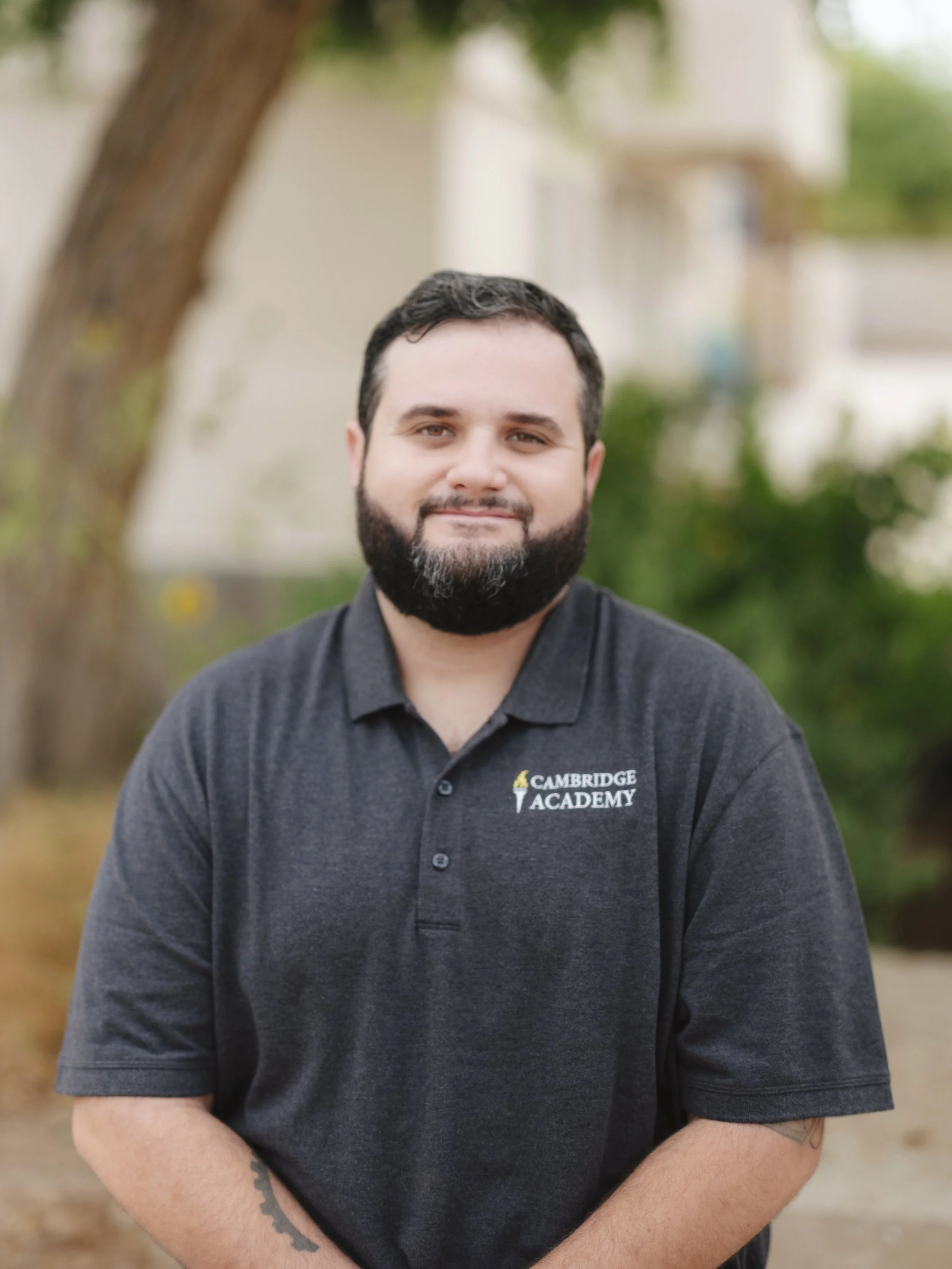 A man with dark hair and a beard, wearing a black polo shirt with the logo of Cambridge Academy, standing outdoors with blurred trees and buildings in the background.