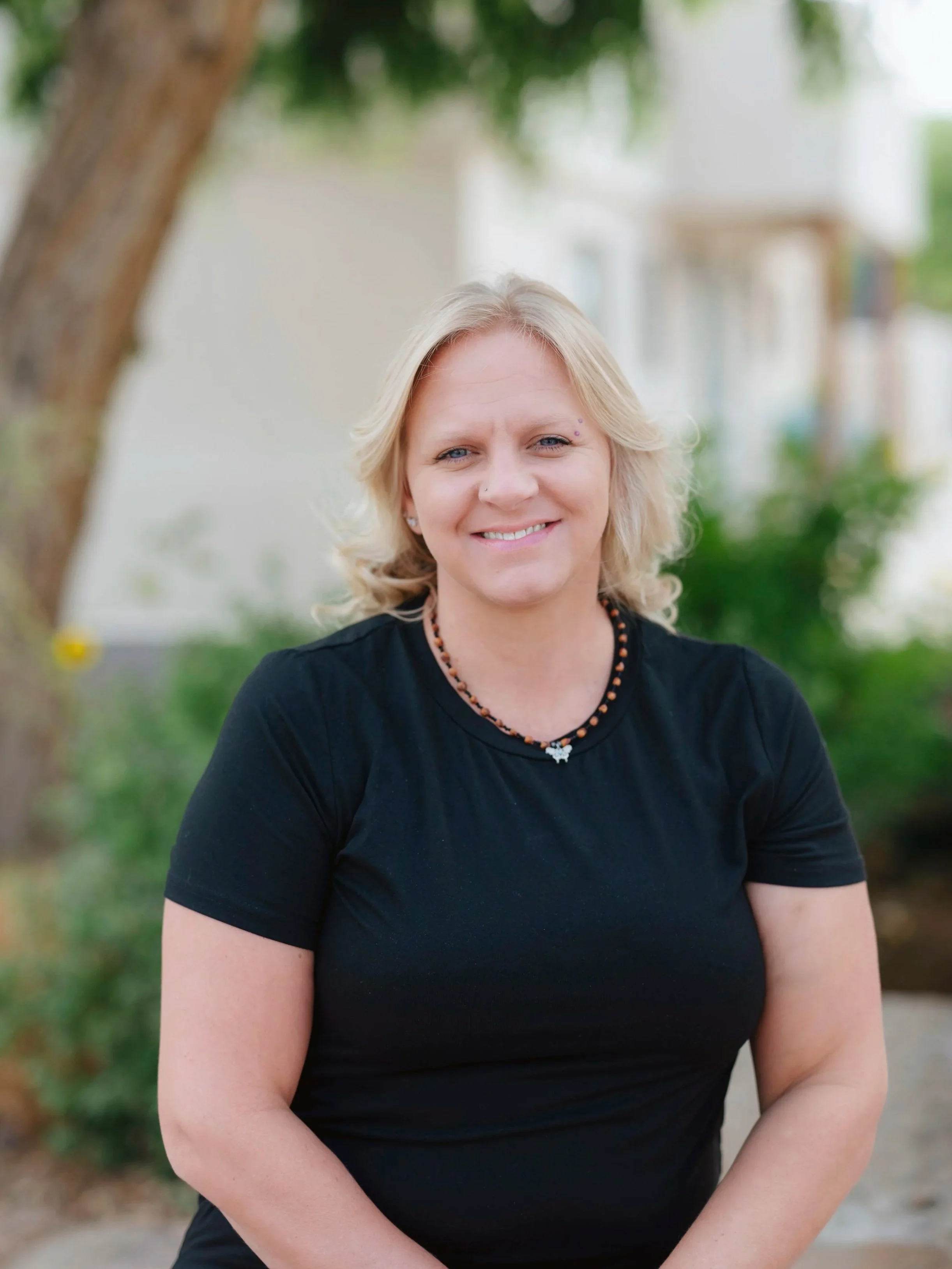 A woman with short blonde hair smiling, wearing a black shirt and a beaded necklace, standing outdoors with greenery and a building in the background.