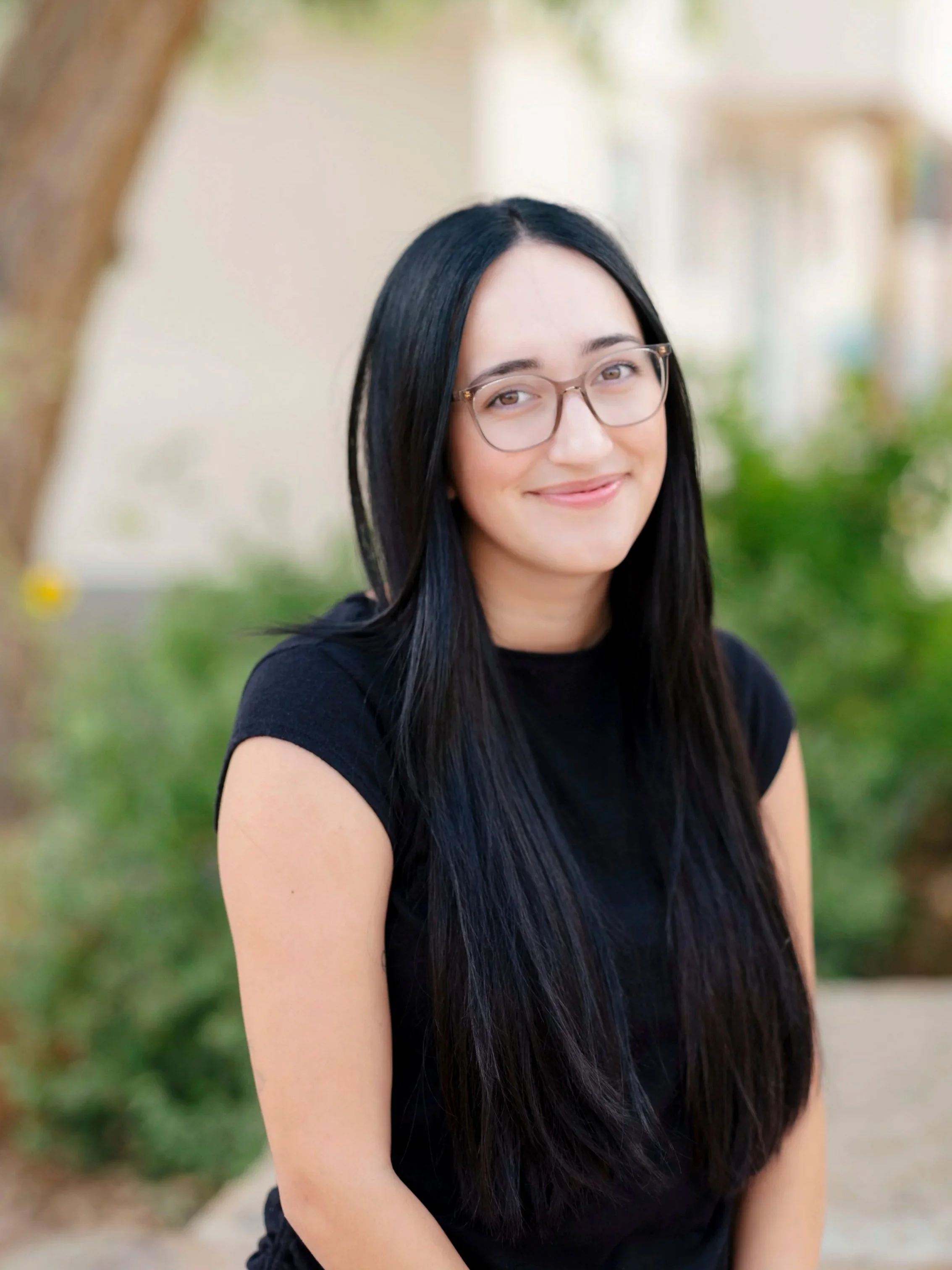 A young woman with long black hair and glasses smiling outdoors in front of blurred trees and greenery.