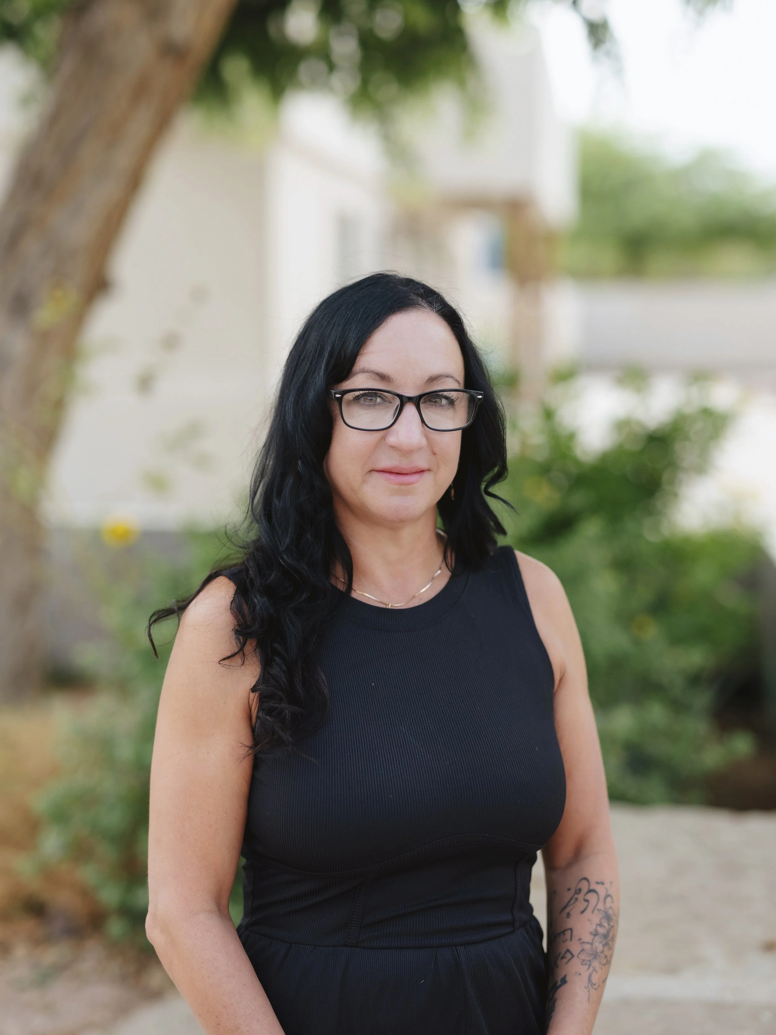A woman with long black hair, glasses, and tattoos on her right arm, wearing a black sleeveless top, standing outdoors with trees and greenery in the background.