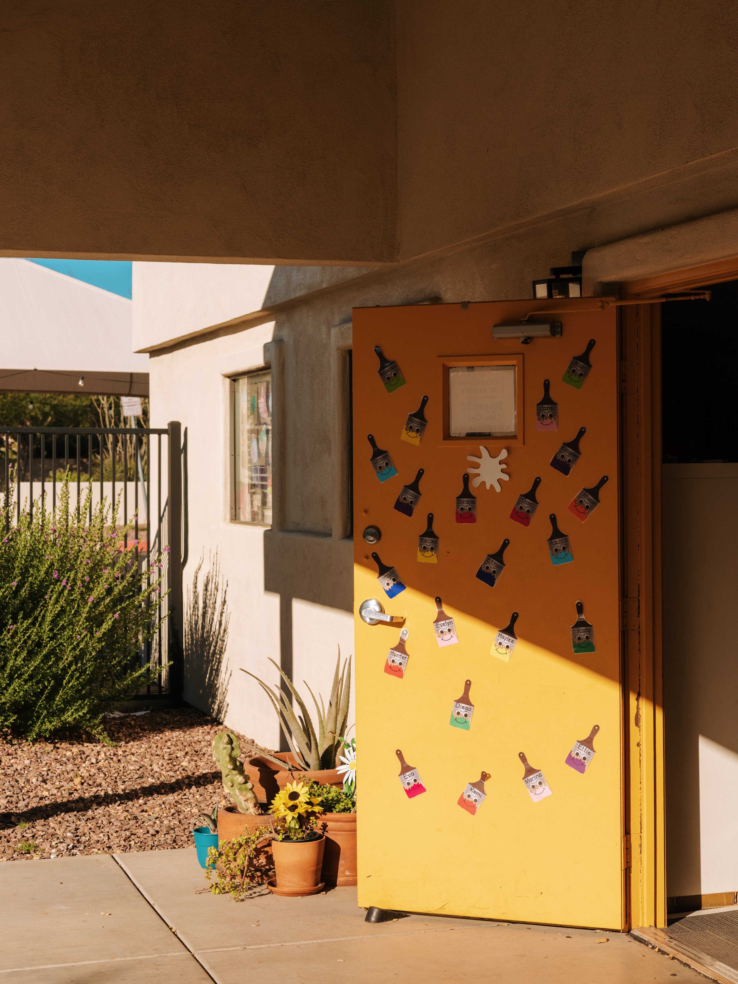Two yellow doors with small windows above; a window with a 'Welcome!' sign and decorative bee figures; potted plants on the ground; string lights hanging above.