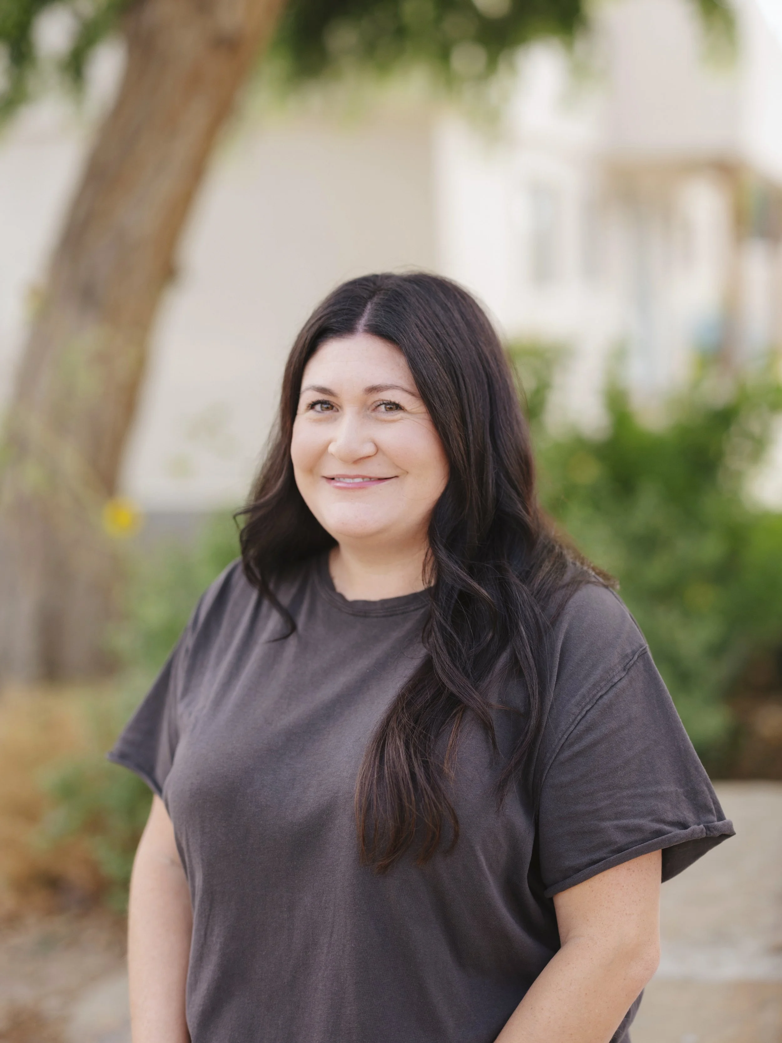 A woman with long dark hair and a dark shirt smiling outdoors near a tree with a blurred background.