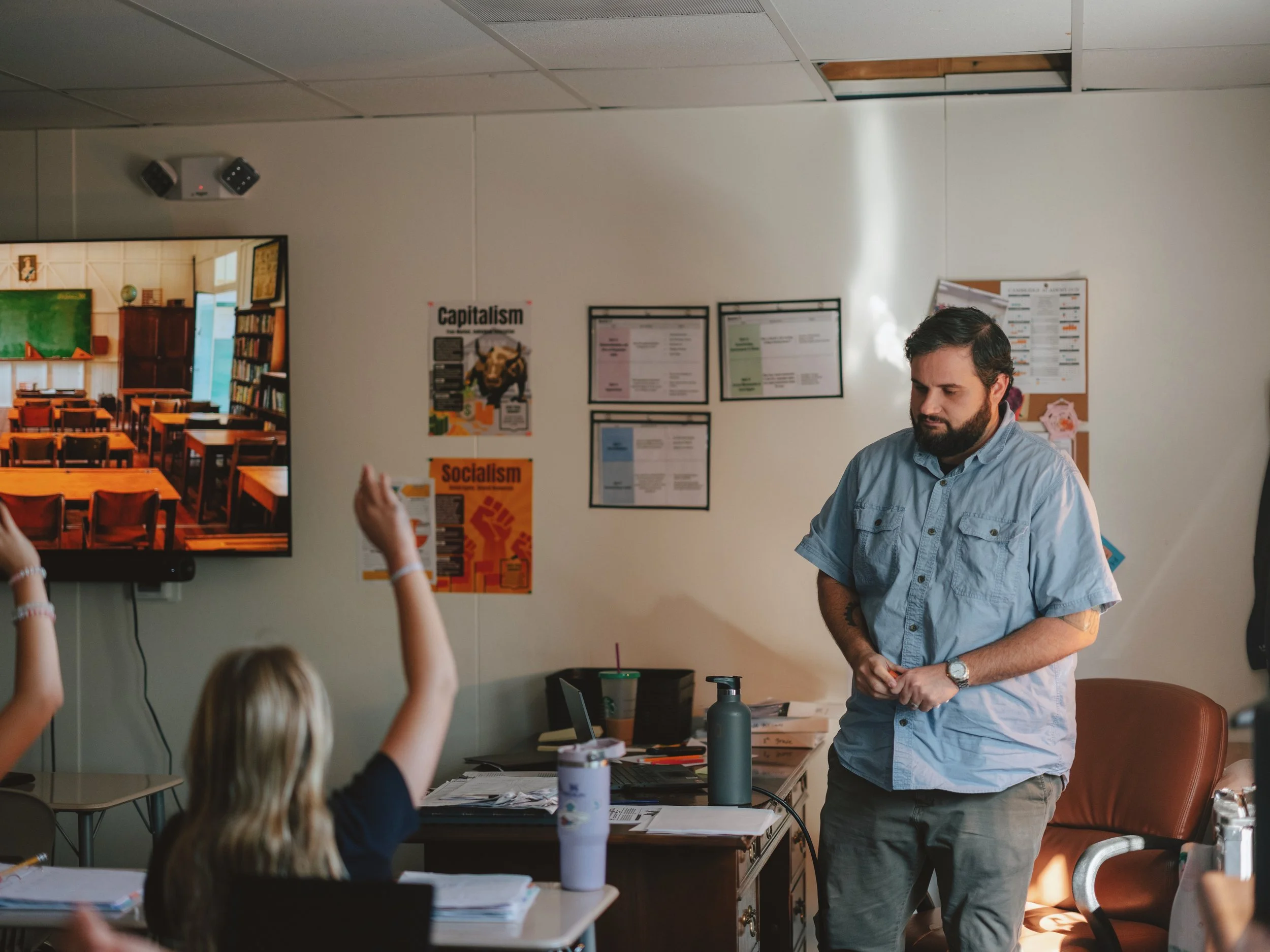 A classroom scene with students raising their hands, and a teacher standing near a desk with books and papers, watching the students.