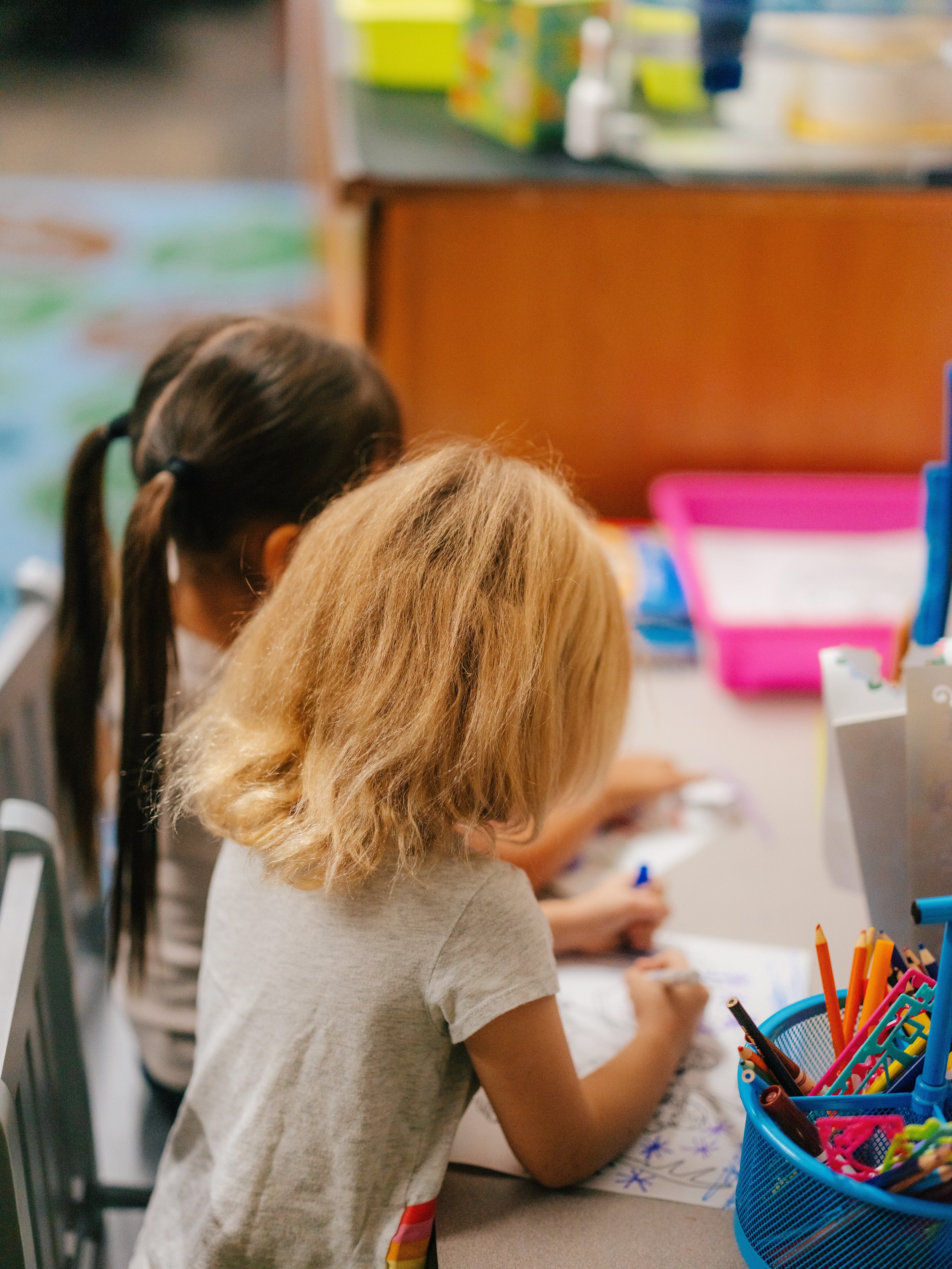 Two young girls, one with blonde hair and another with dark hair in pigtails, are sitting at a desk coloring with markers and colored pencils, with art supplies and storage bins visible.