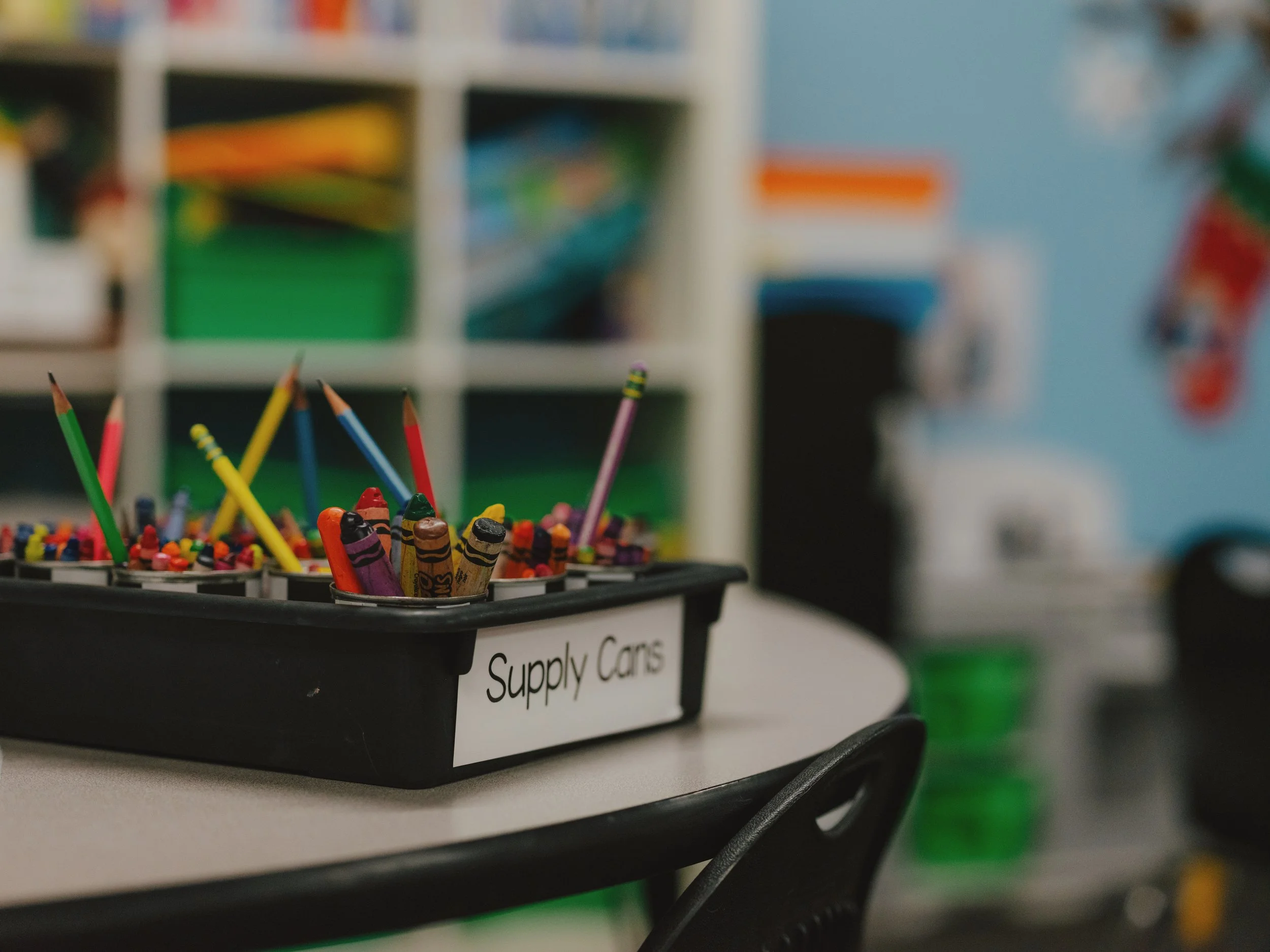 A young girl in a teal shirt is smiling and adding a test tube to a rack of test tubes, while an adult stands next to her observing in a laboratory or classroom setting.