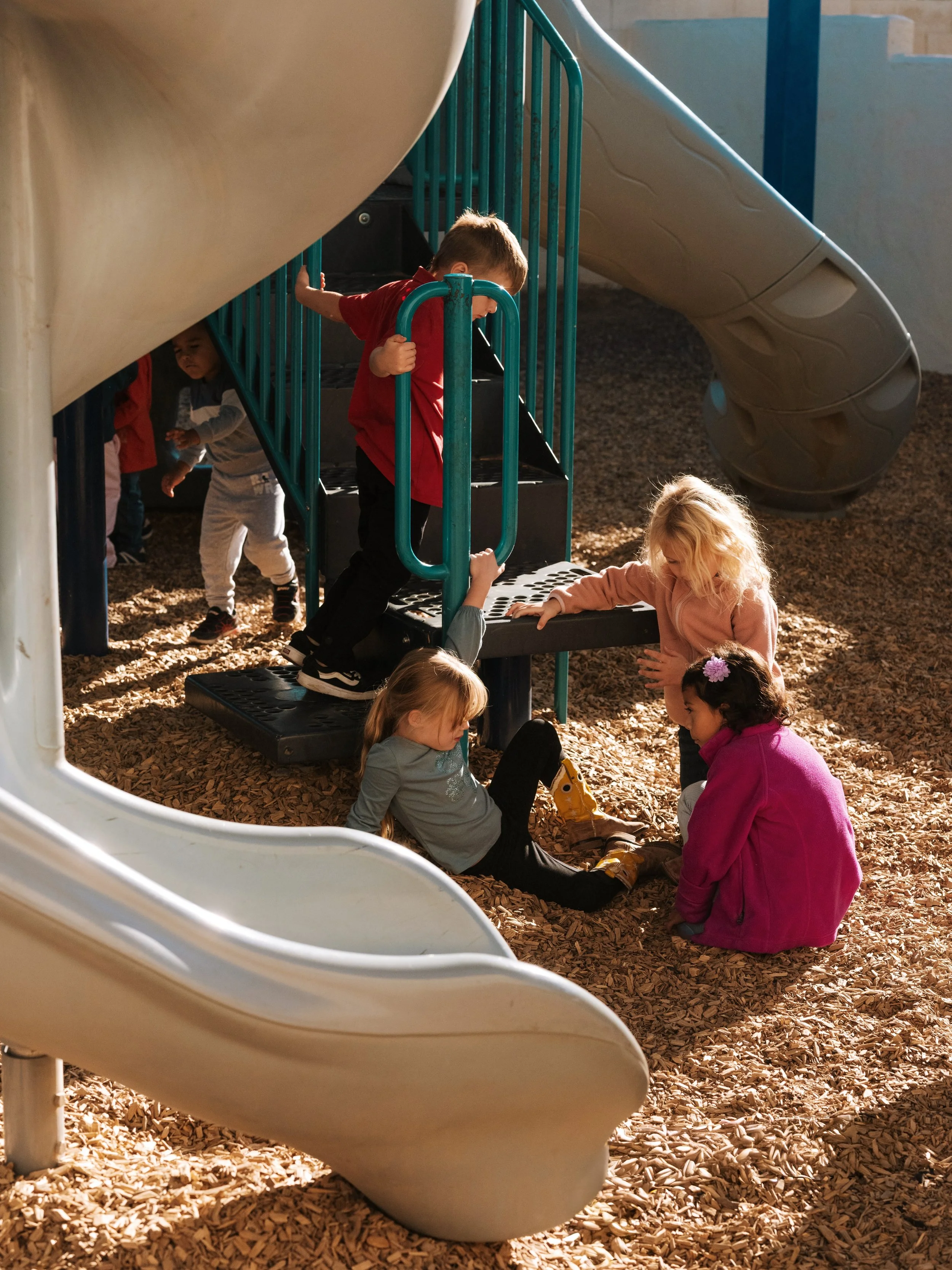 Children playing on playground equipment with slides and stairs, with wood chips on the ground.