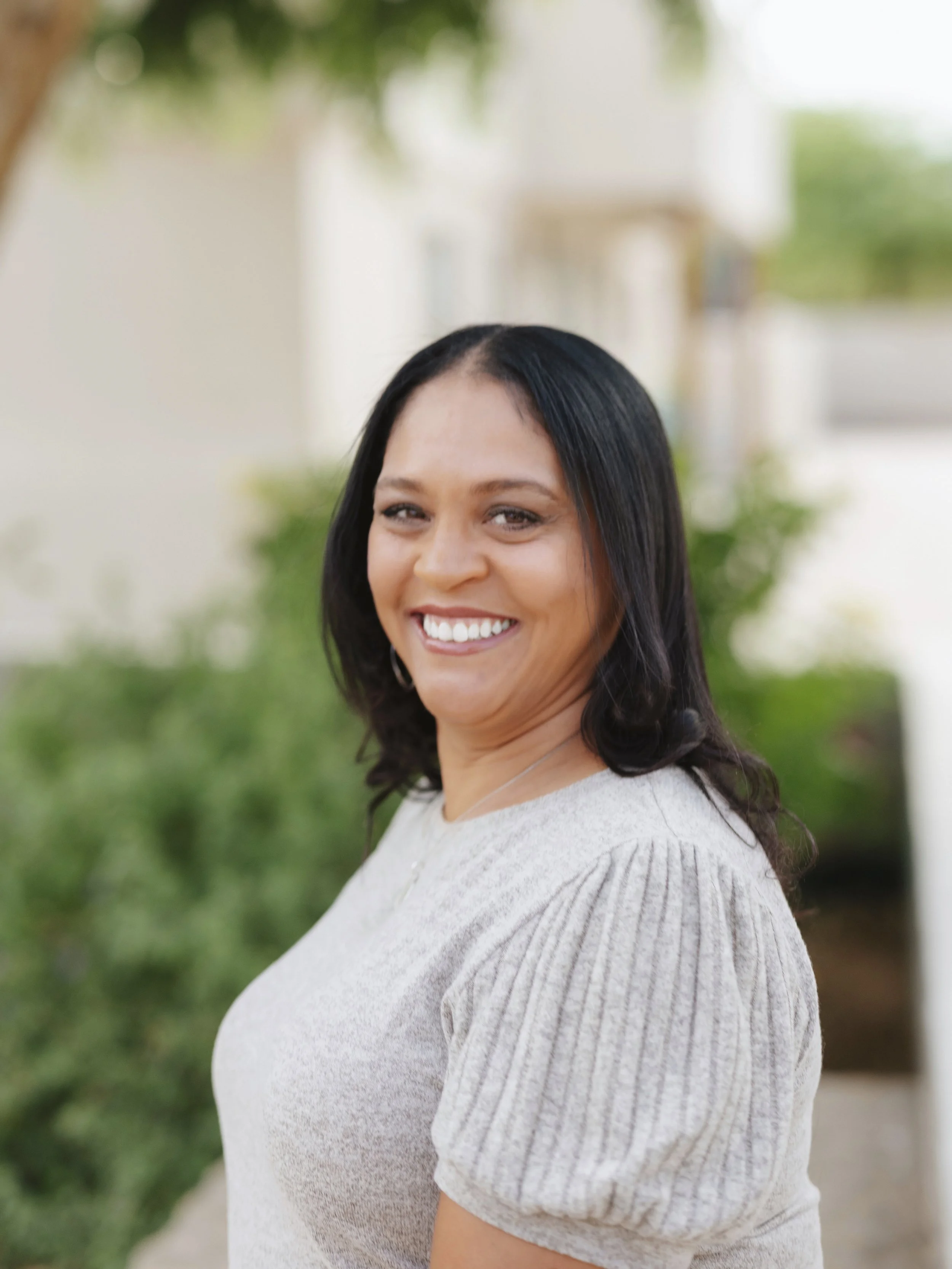A woman with dark hair smiling outdoors with greenery and a white building in the background.