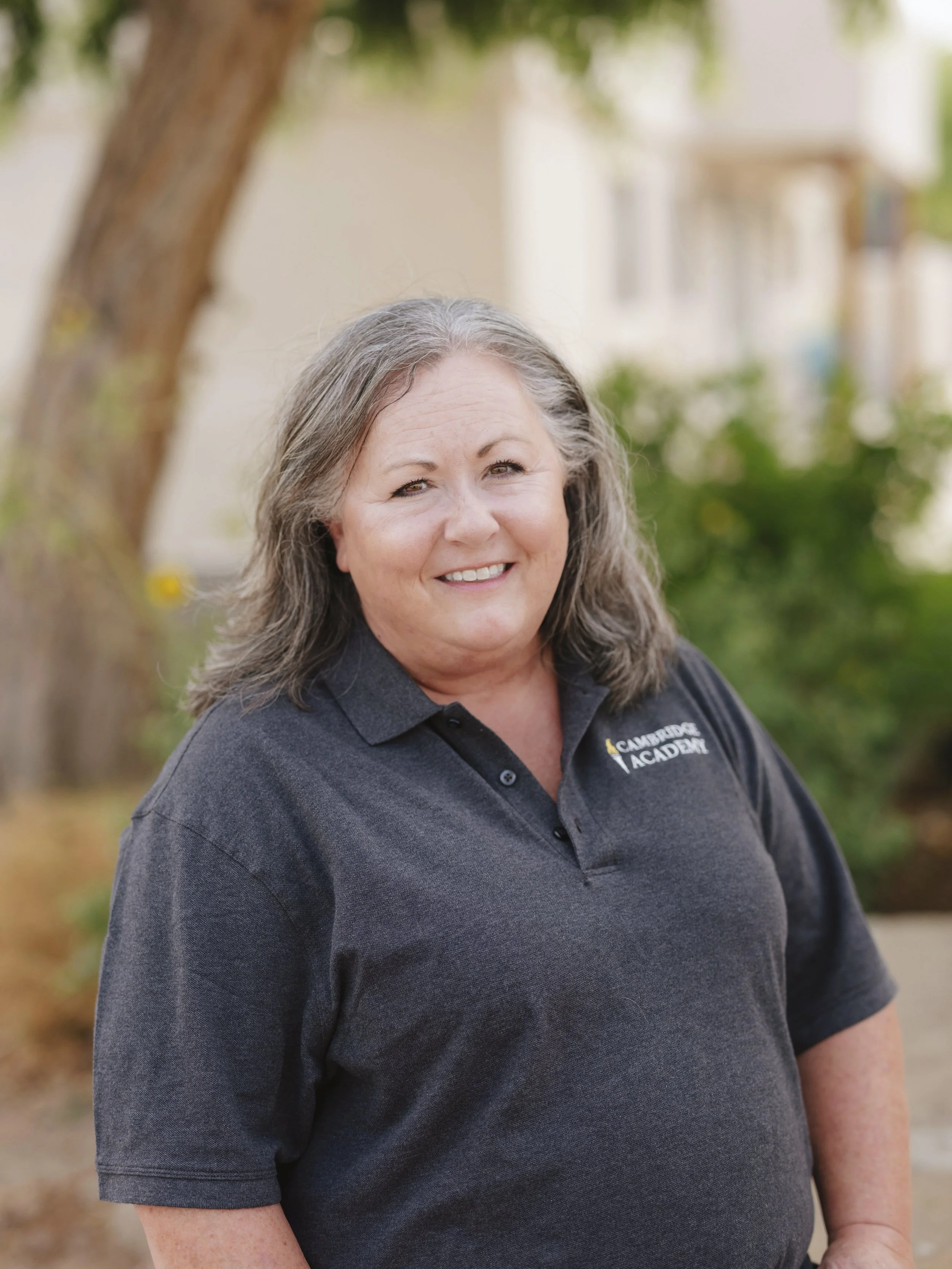 A woman with gray hair smiling outdoors, wearing a dark gray polo shirt with a logo that says 'Cambridge Academy'.