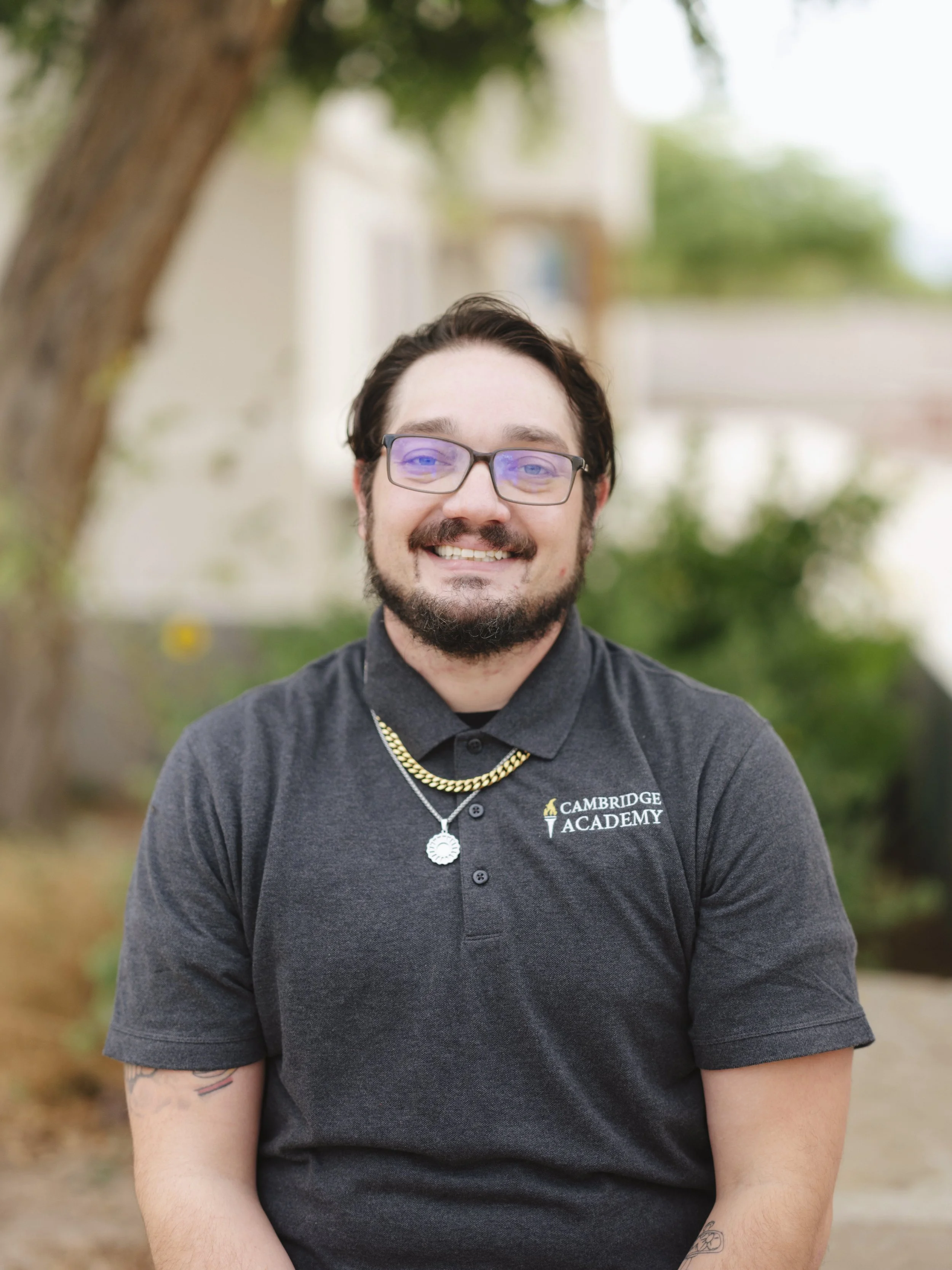 Smiling man with glasses, dark hair, and a beard, wearing a dark gray polo shirt with a logo that says 'Cambridge Academy,' standing outdoors near a tree with blurred green and light-colored background.