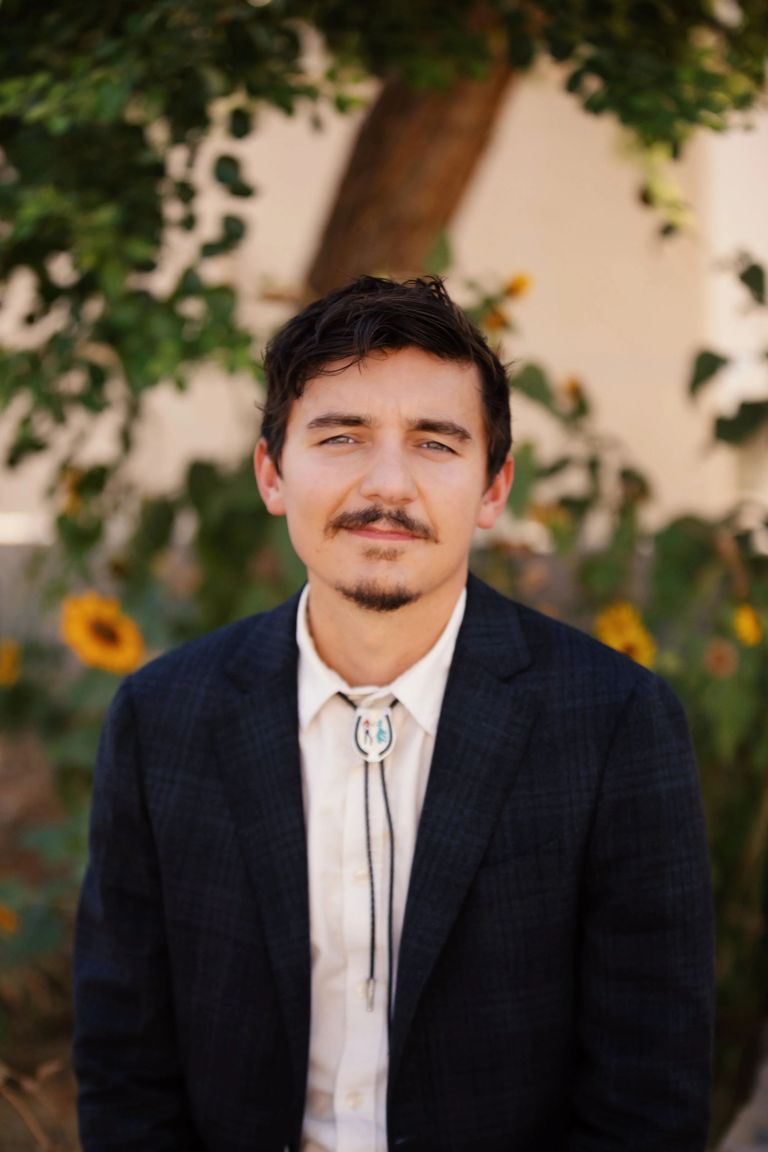 A young man with dark hair, a small mustache, and a goatee, wearing a white shirt with a bolo tie and a dark blazer, standing outdoors in front of a tree and sunflower plants.